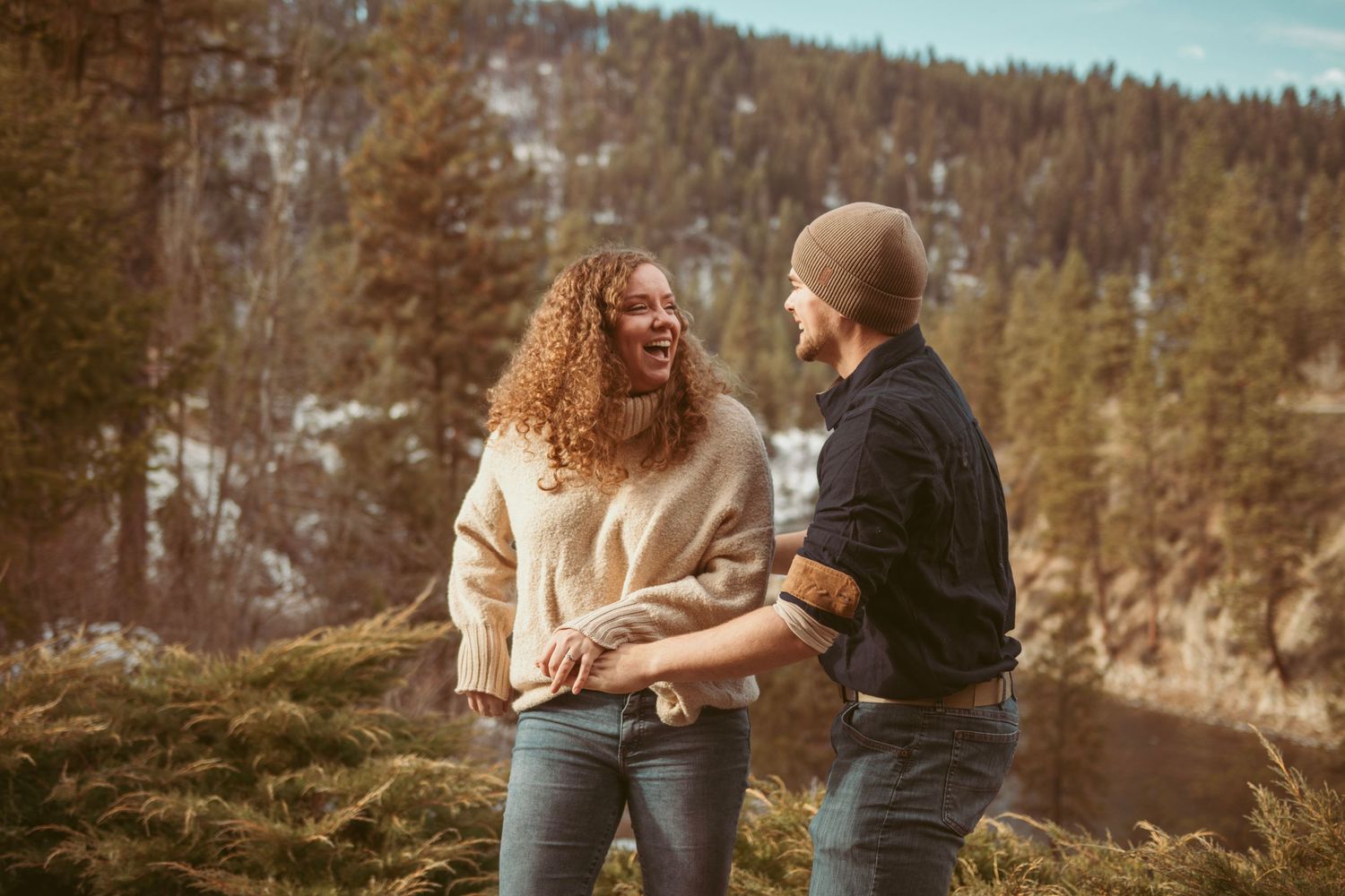 A playful couple shares laughter while sitting on rocky terrain with snow-capped mountains in background.