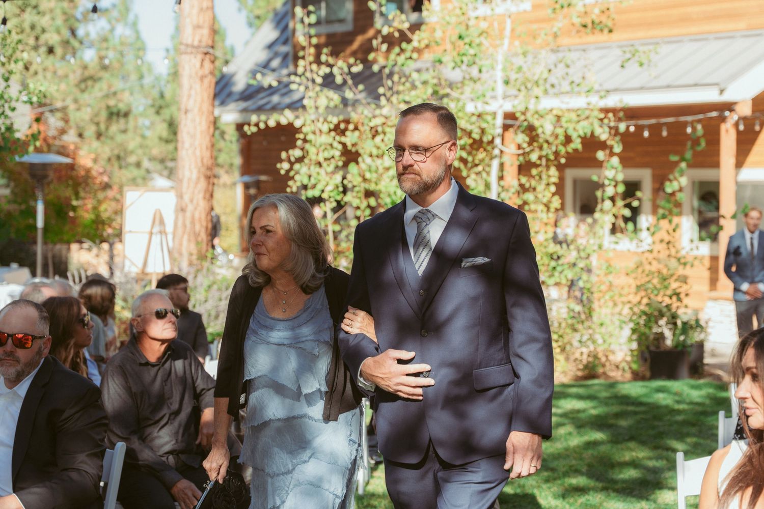 Wedding guests walk down an aisle at an outdoor ceremony with string lights and trees in the background.