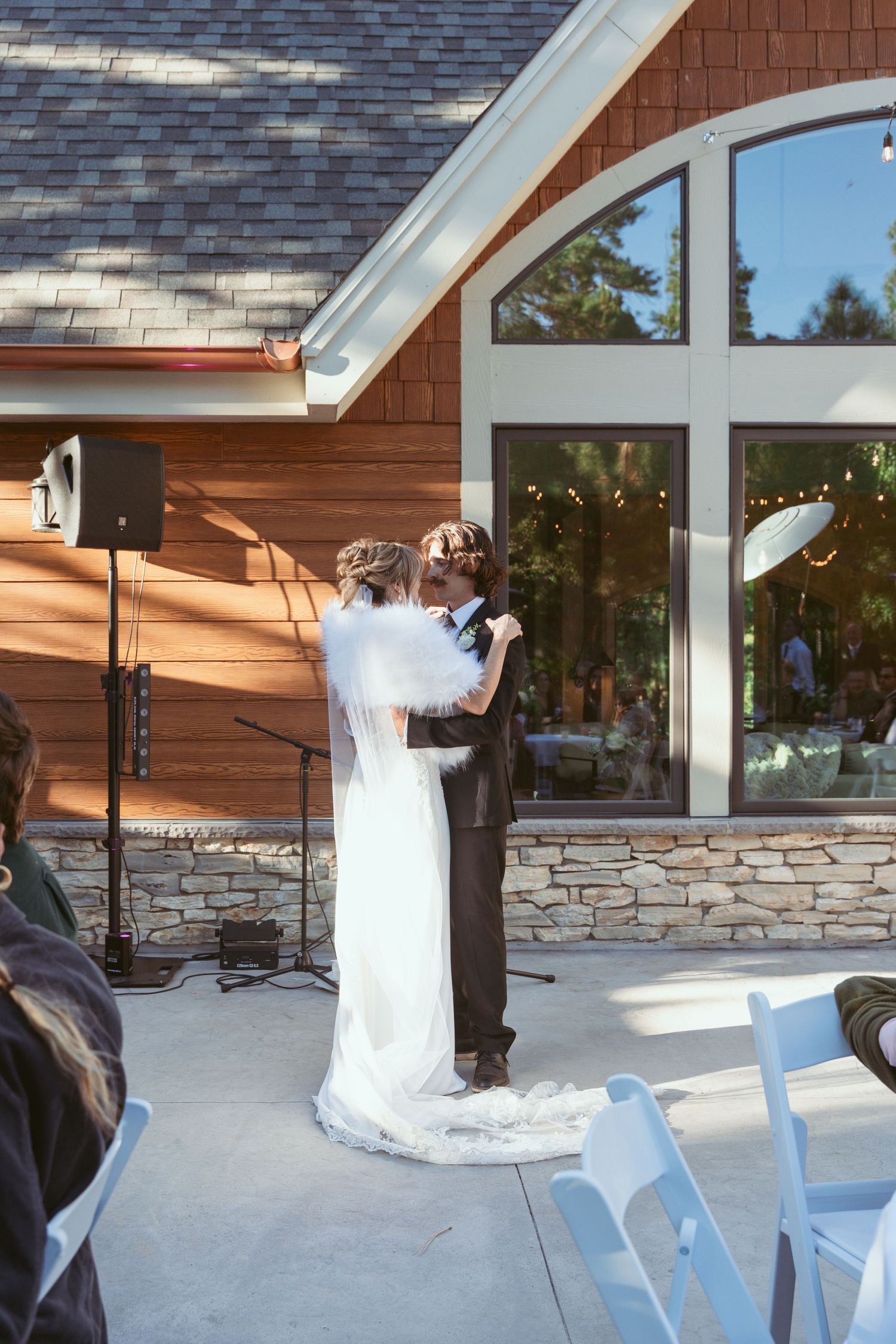 A couple shares their first dance at an outdoor wedding venue with stone walls and large windows.