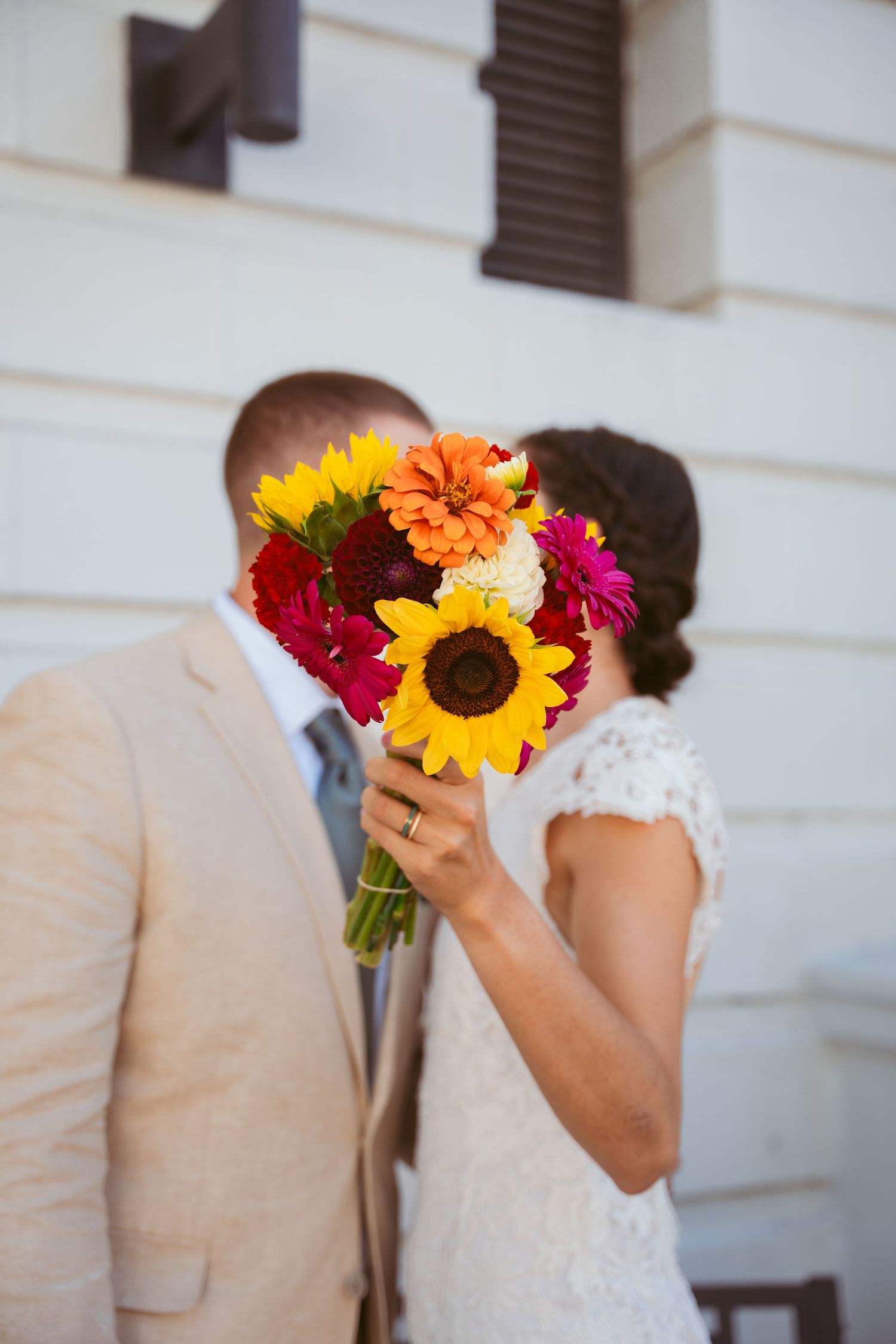 Couple holding vibrant sunflower and wildflower bouquet against white building wall.