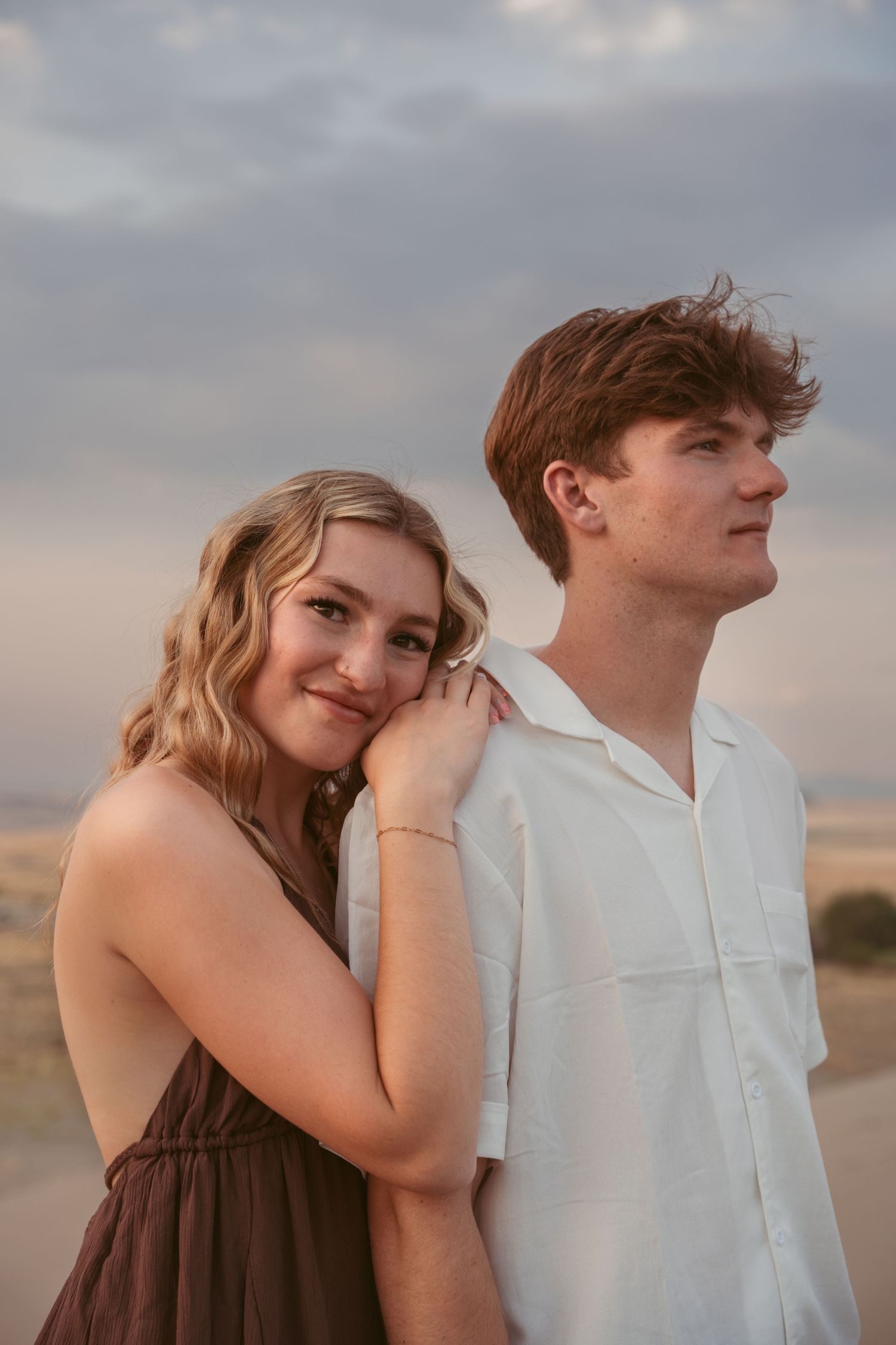 A couple in brown dress and white shirt share tender moments on desert dunes at sunset.