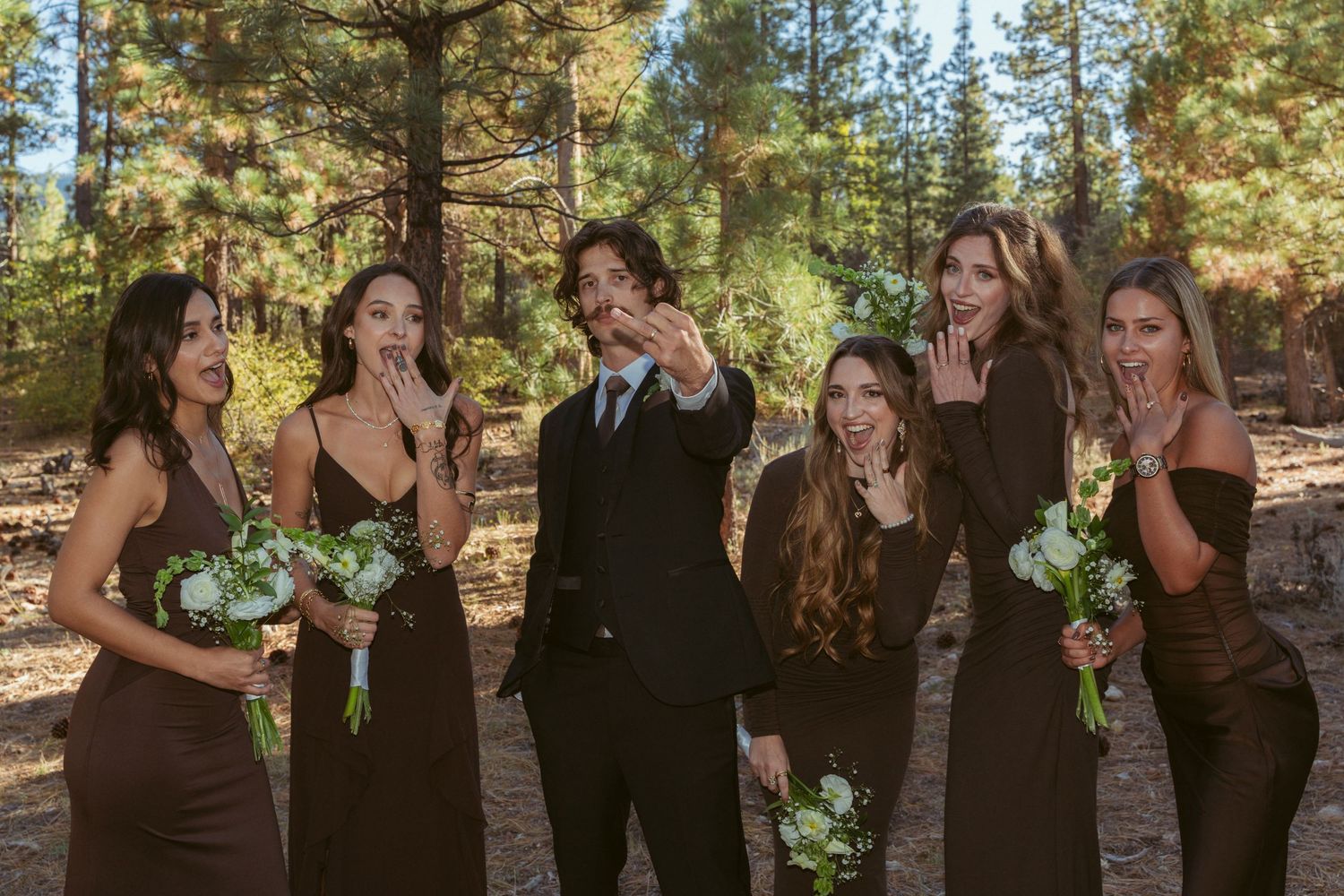 Wedding party members lined up outdoors among pine trees wearing formal attire.