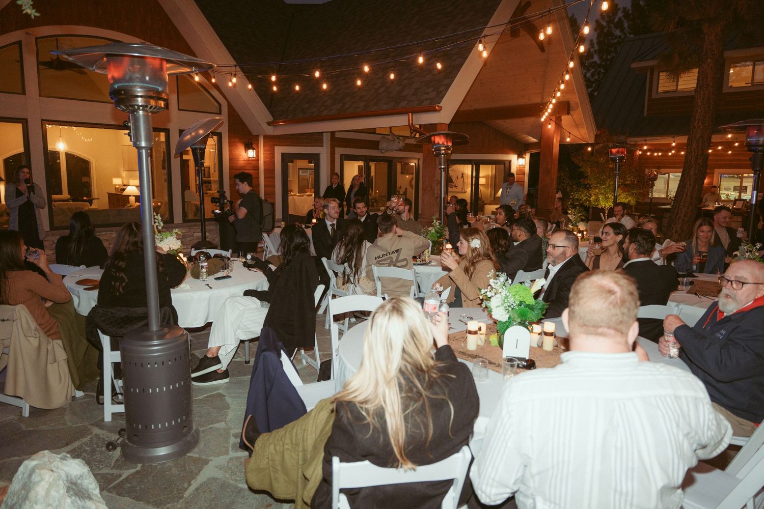 Wedding guests gathered at reception tables under warm lighting in an outdoor evening celebration.