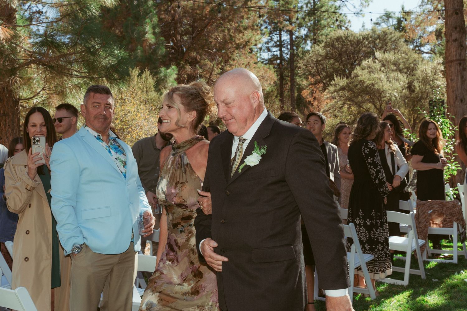 Wedding guests interacting during outdoor ceremony beneath pine trees on grassy venue.