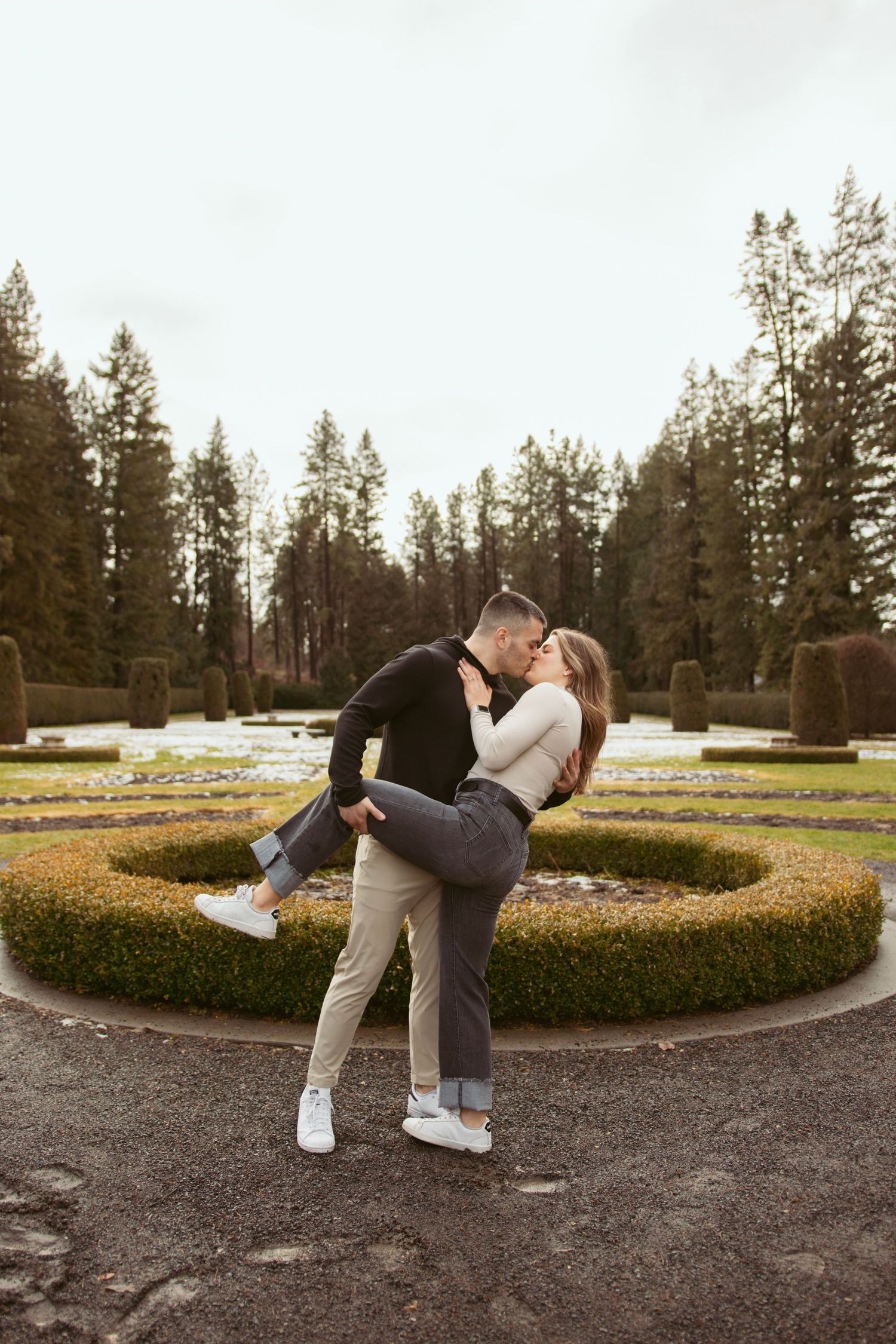 A couple shares romantic poses in a garden with evergreen trees and manicured hedges.