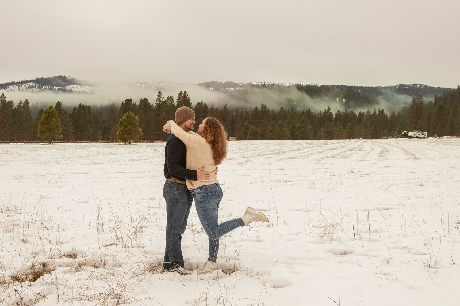 Wide shot of couple embracing in snowy field with mountains in background.