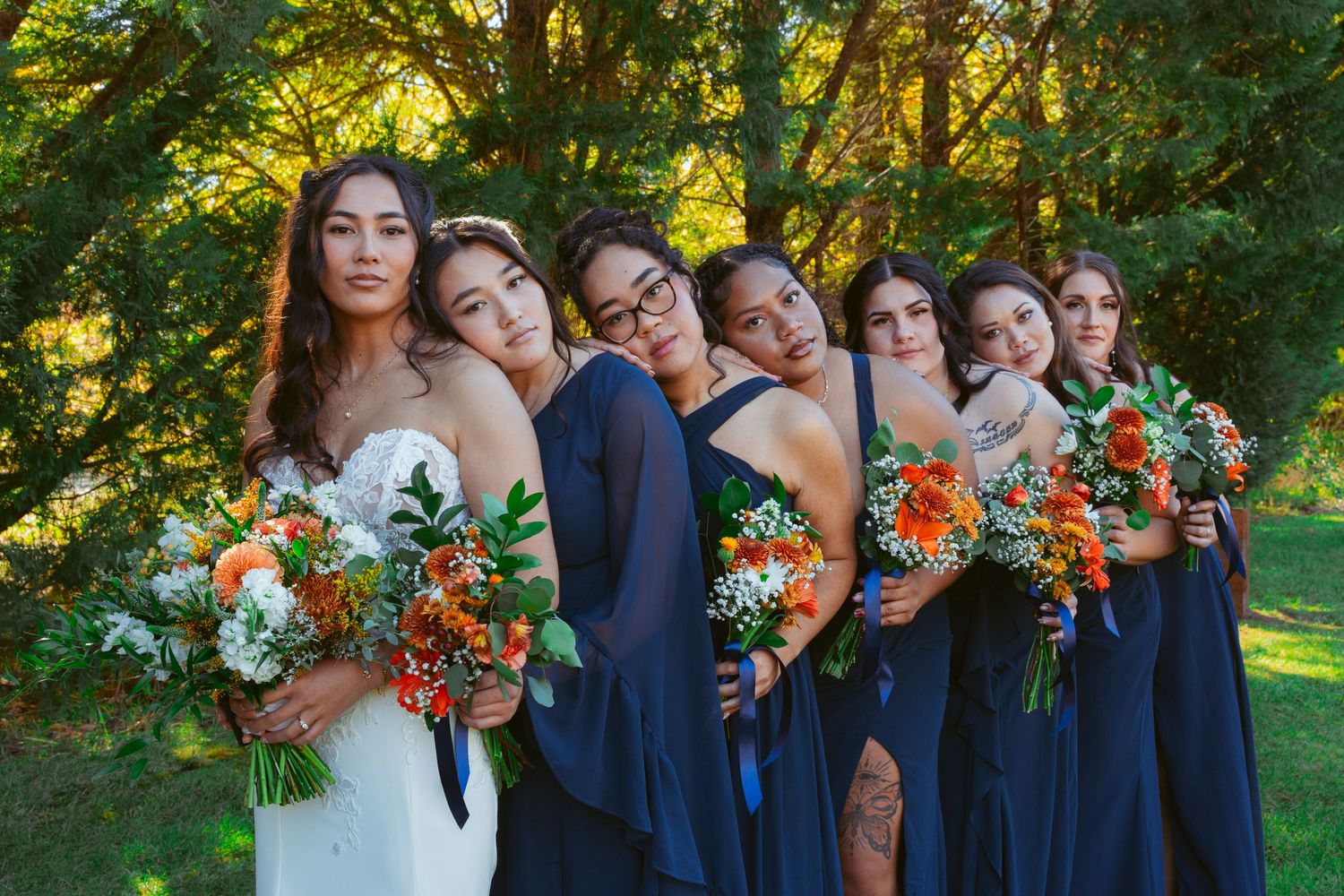 Wedding party poses together outdoors with navy blue dresses and bright orange bouquets against green foliage.
