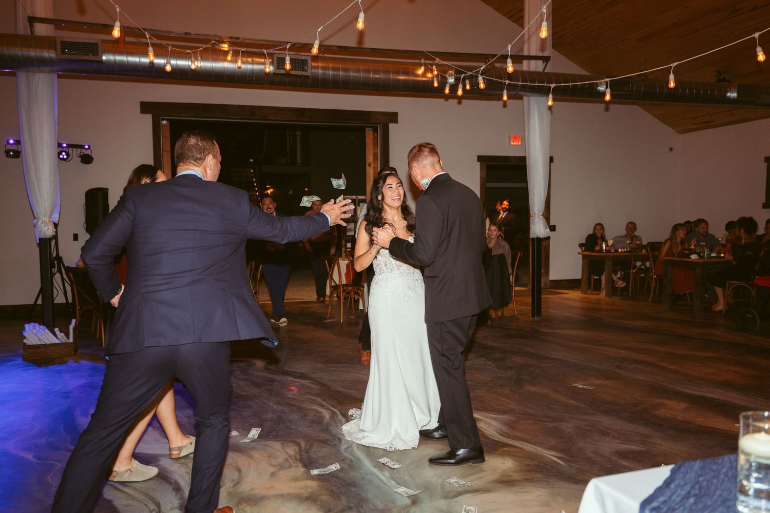 Wedding celebration on a wooden dance floor with decorative string lights and white walls.
