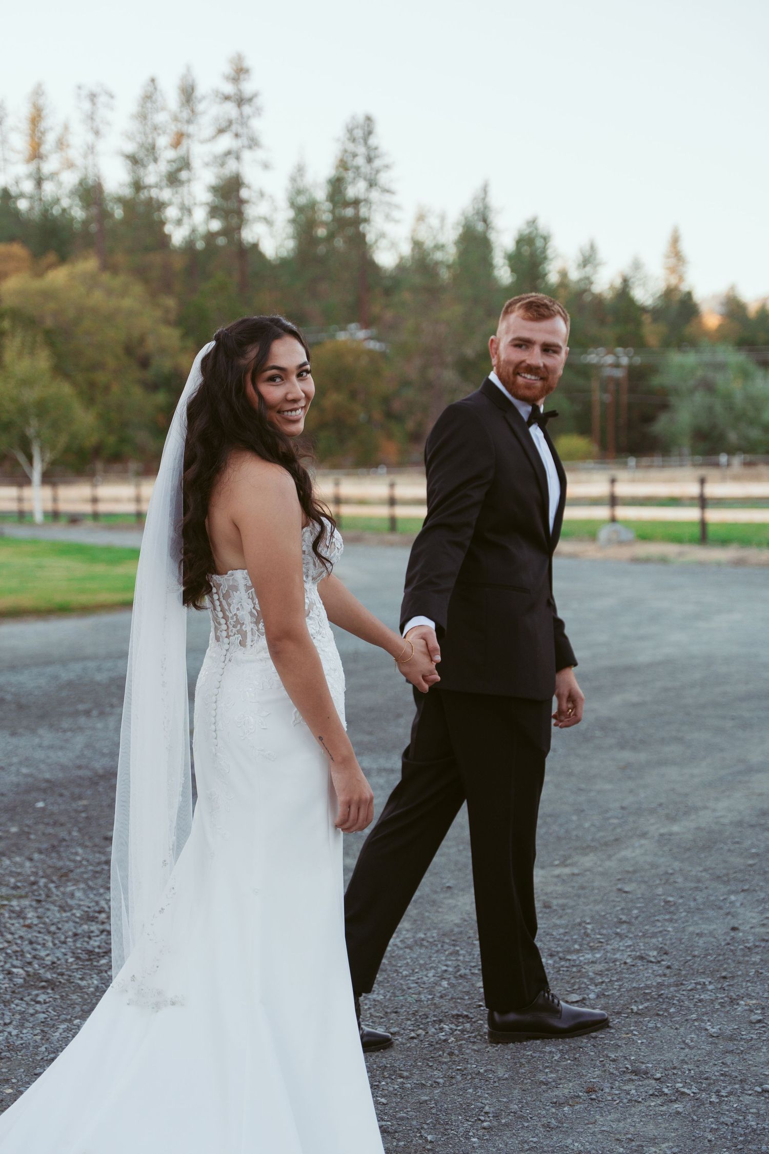 Wedding couple walks together on a gravel path with evergreen trees in the background.