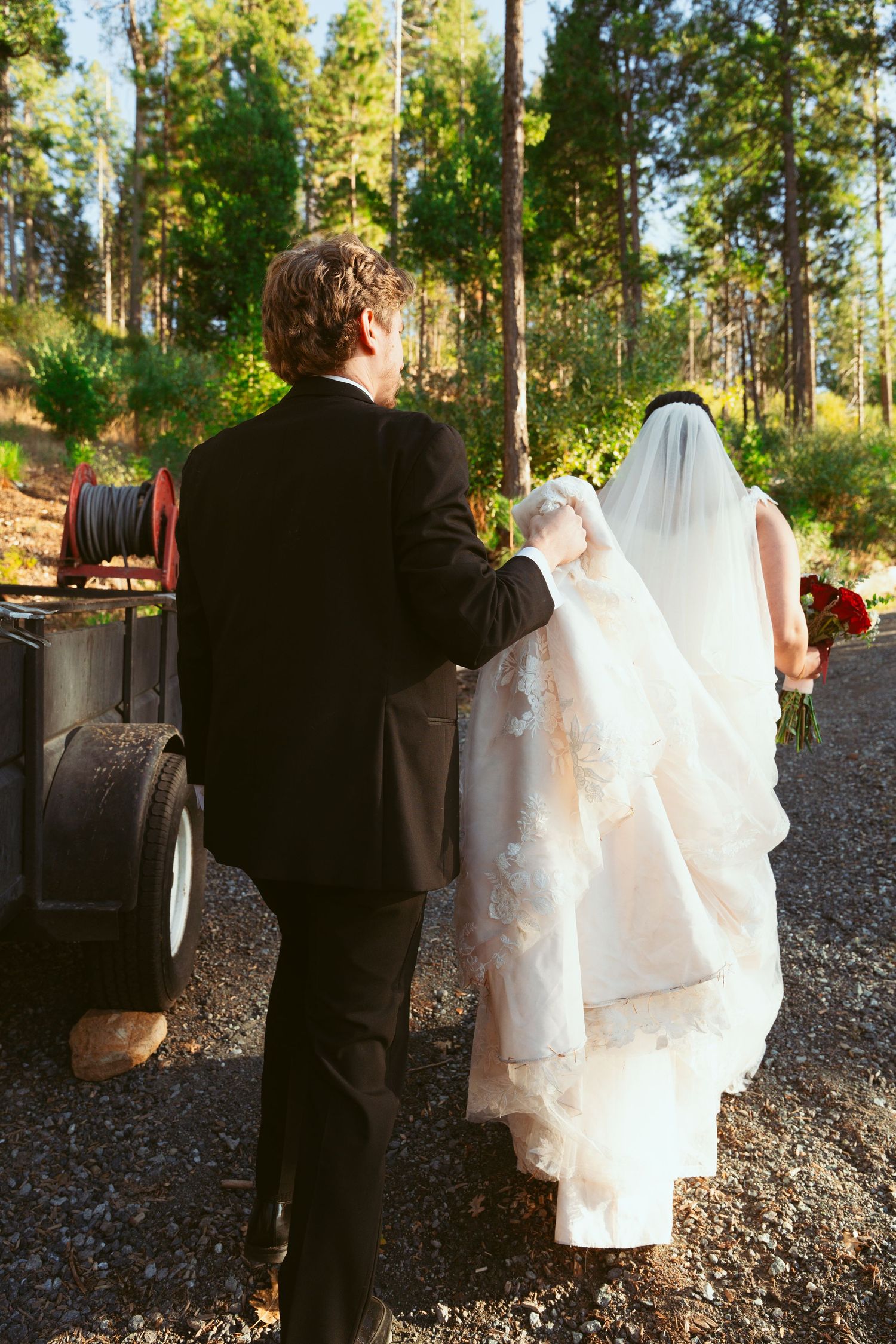 A couple in wedding attire walks through a wooded outdoor setting with tall pine trees.