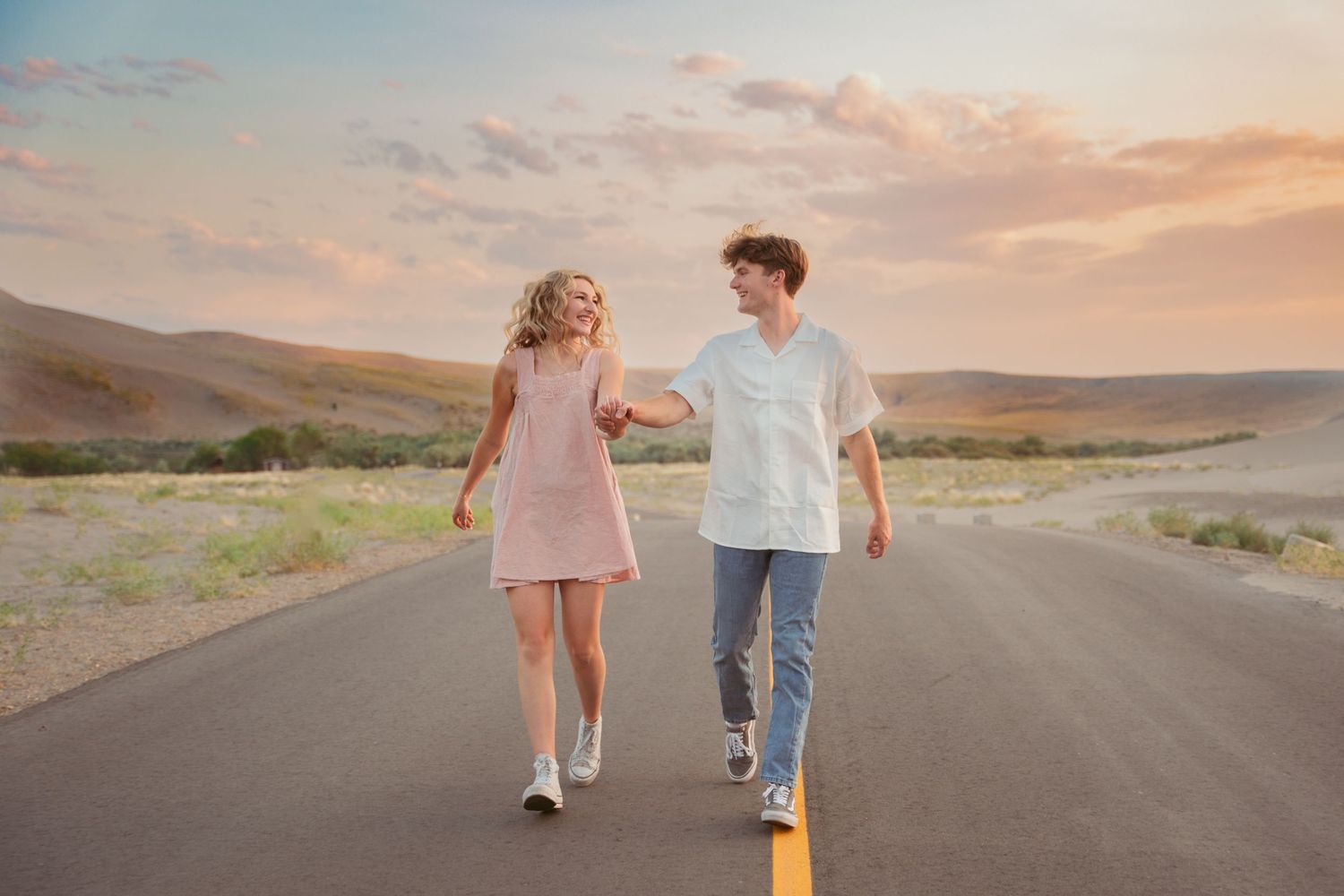 A couple walks hand in hand down an empty desert road during golden hour.