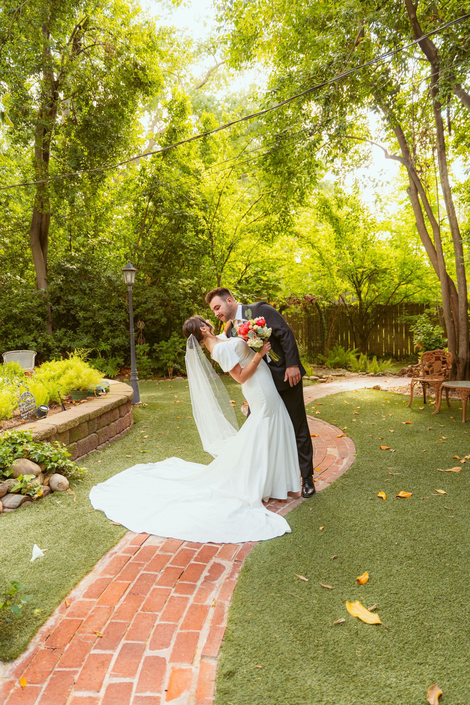 Romantic dip kiss between newlyweds on brick pathway surrounded by lush green garden.