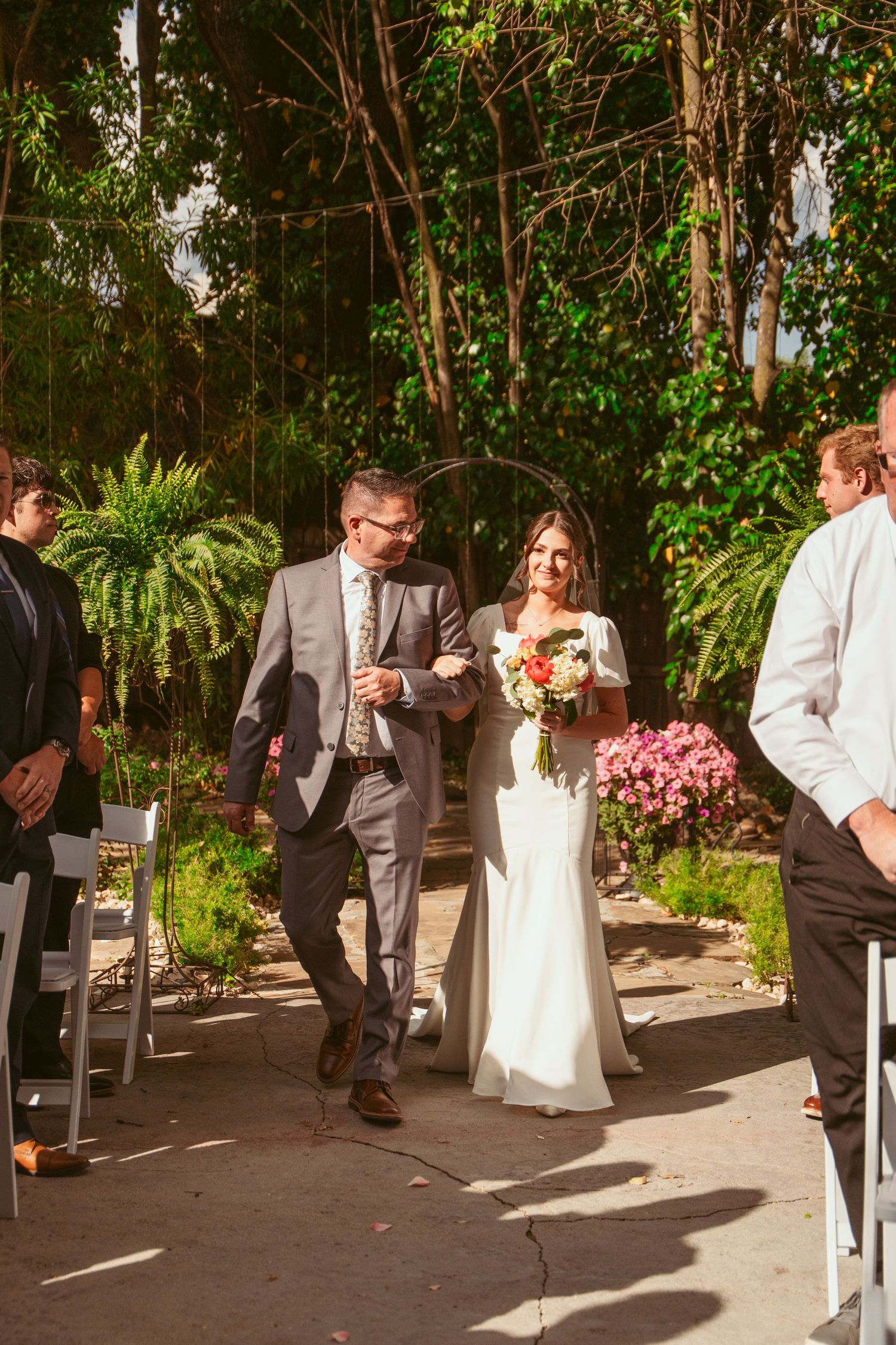 Wedding party walks down outdoor aisle lined with chairs and lush greenery.