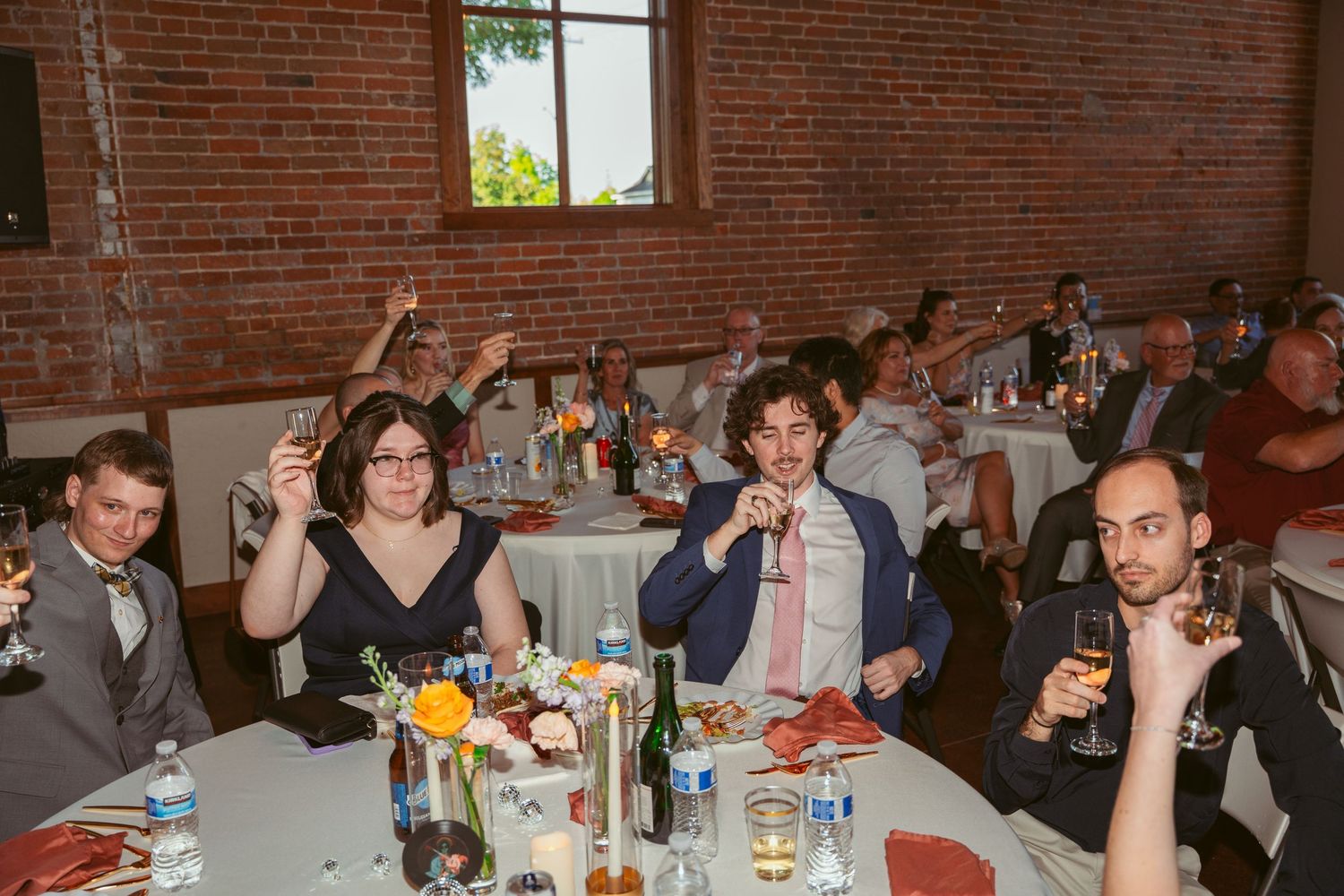 Wedding guests raising their glasses for a toast at round tables during the reception.