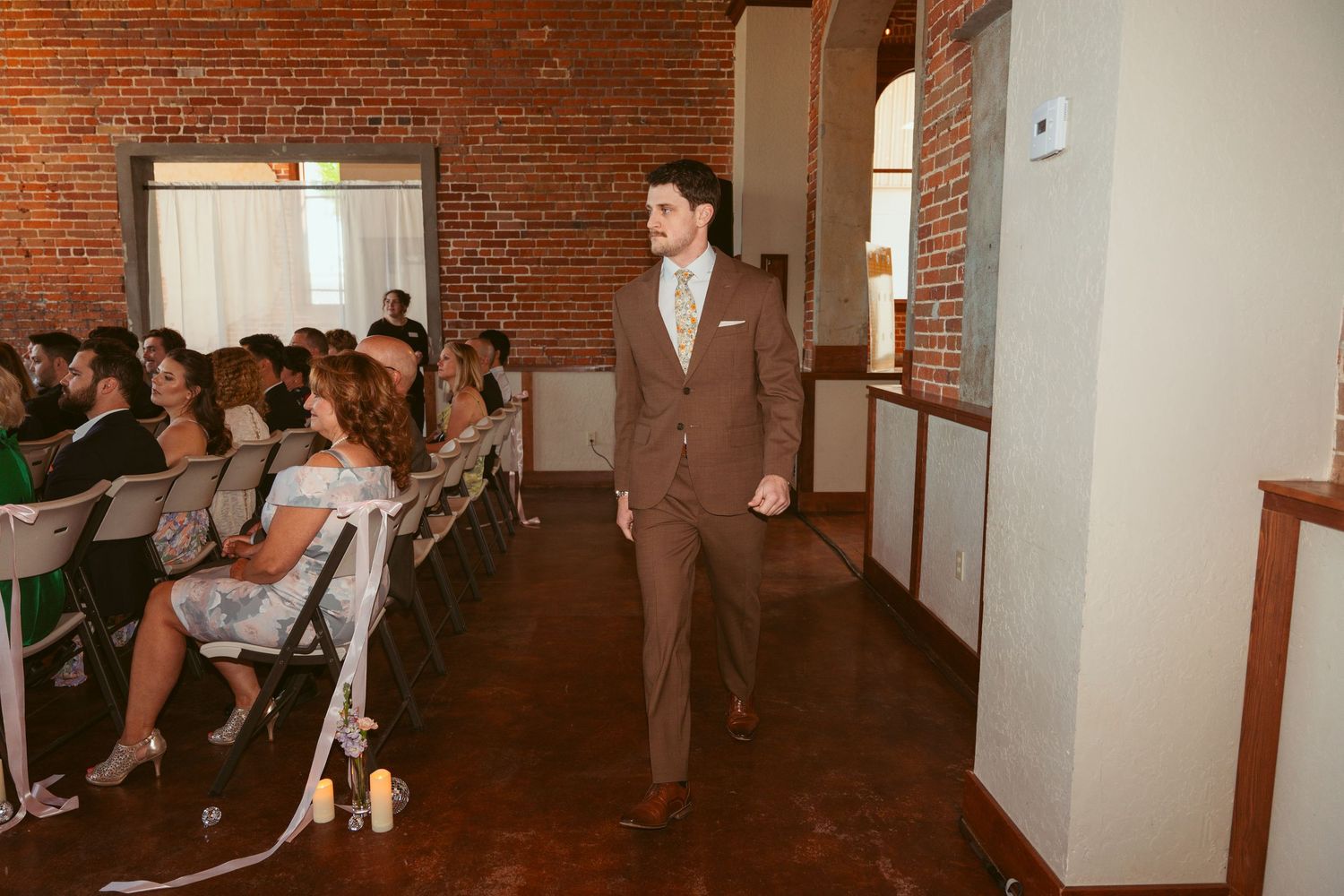 Wedding party member walking down aisle in brown suit at brick-walled ceremony venue.