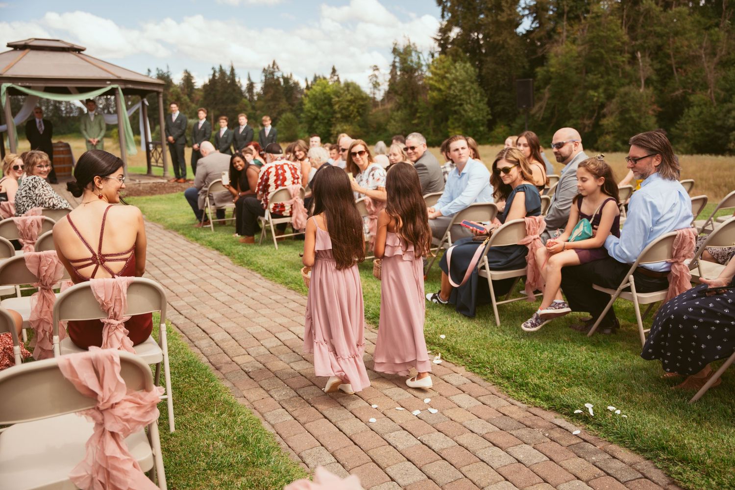 Wedding ceremony setup at an outdoor venue with guests seated on chairs and pink-clad bridesmaids walking down the aisle.