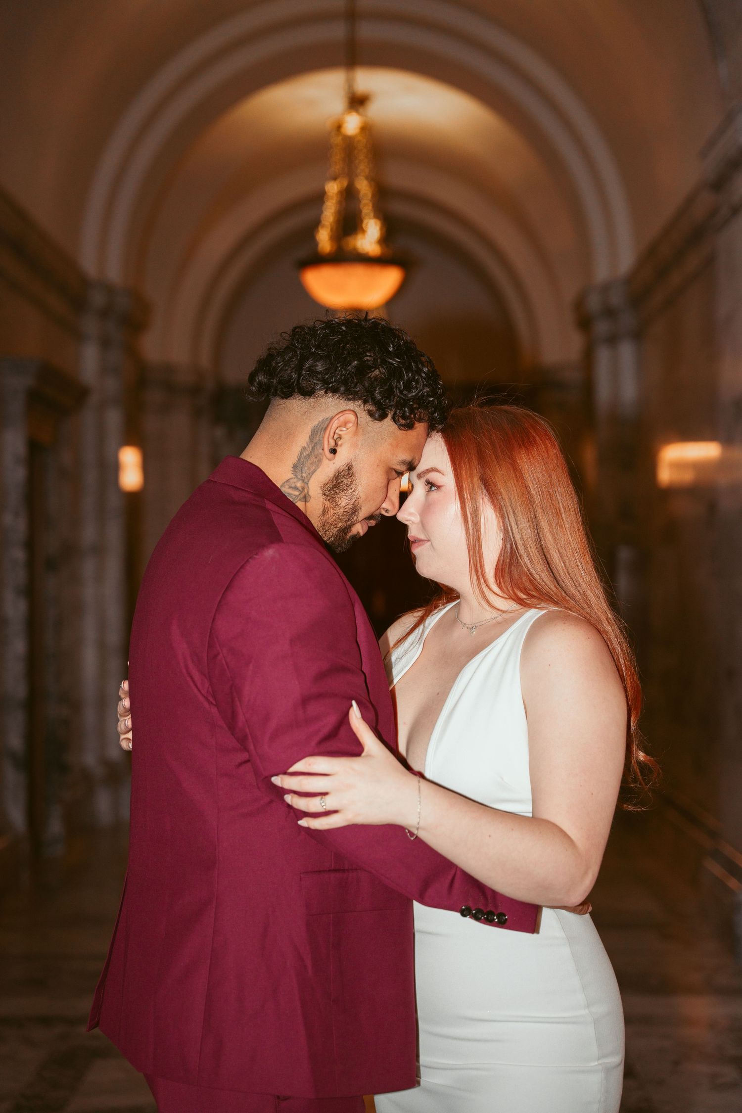 A couple in burgundy and white embraces intimately in a warmly lit historic hallway.