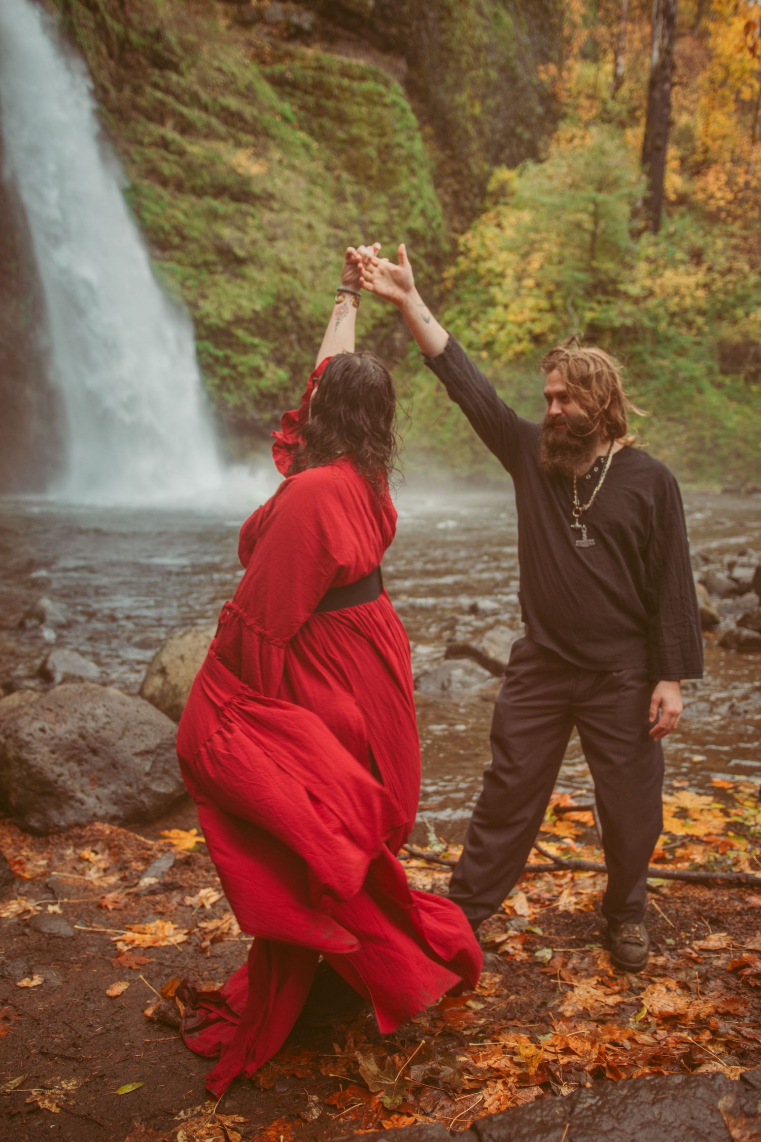 Two people in contrasting red and black outfits pose dramatically near a waterfall surrounded by autumn leaves.