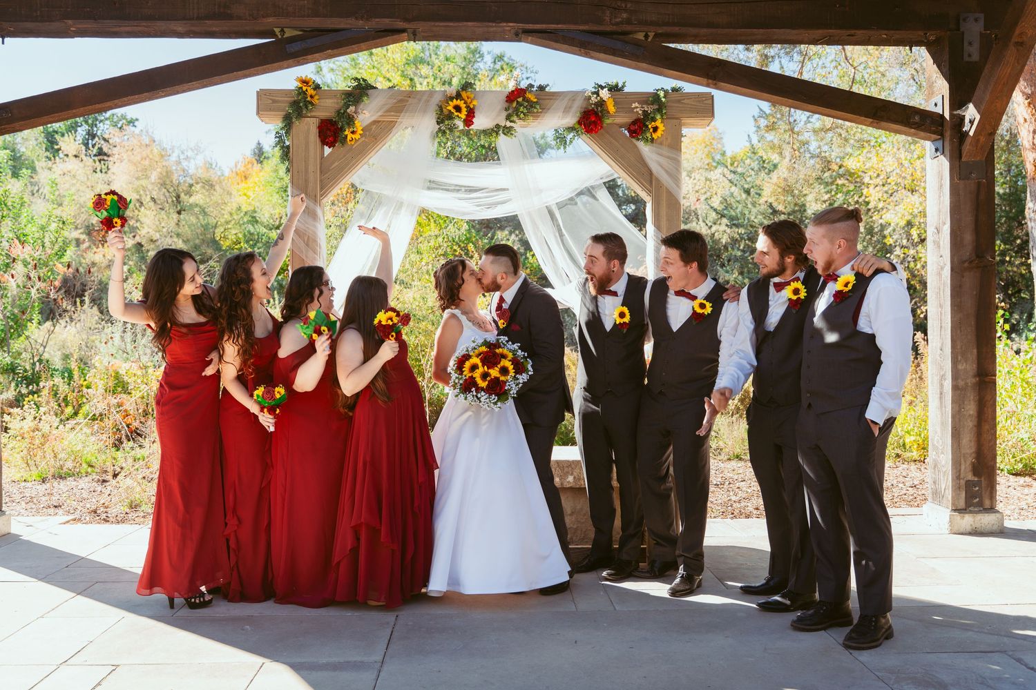 Panoramic shot of entire wedding party posed under wooden pavilion with formal attire.