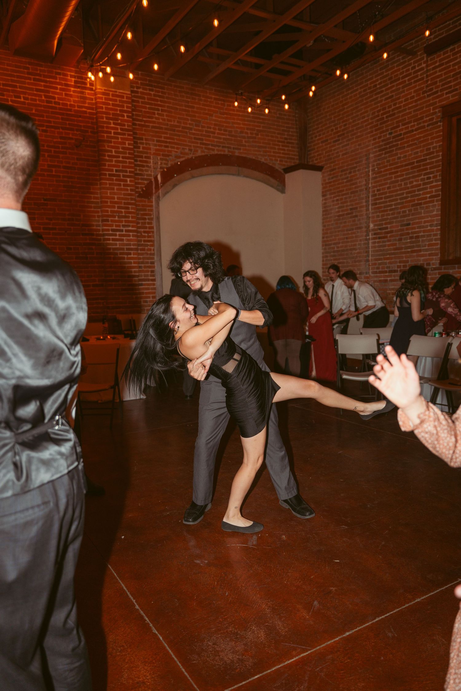 Dancers performing a dramatic dip move on a dance floor under string lights in a brick-walled venue.