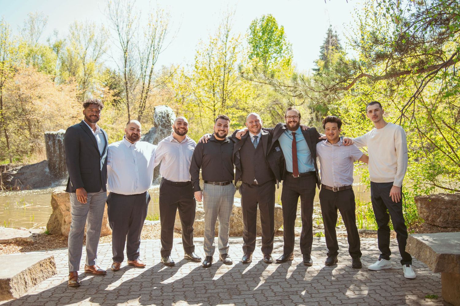 Wedding party members stand in line against a scenic outdoor backdrop with trees and greenery.