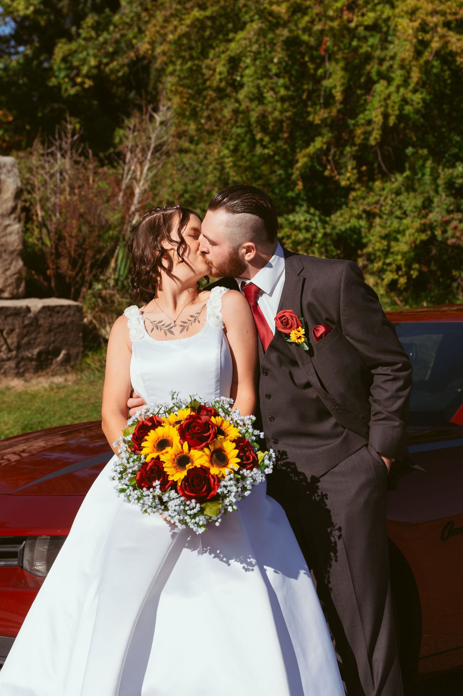 Wedding couple shares romantic moment by vintage red car with sunflower bouquet.