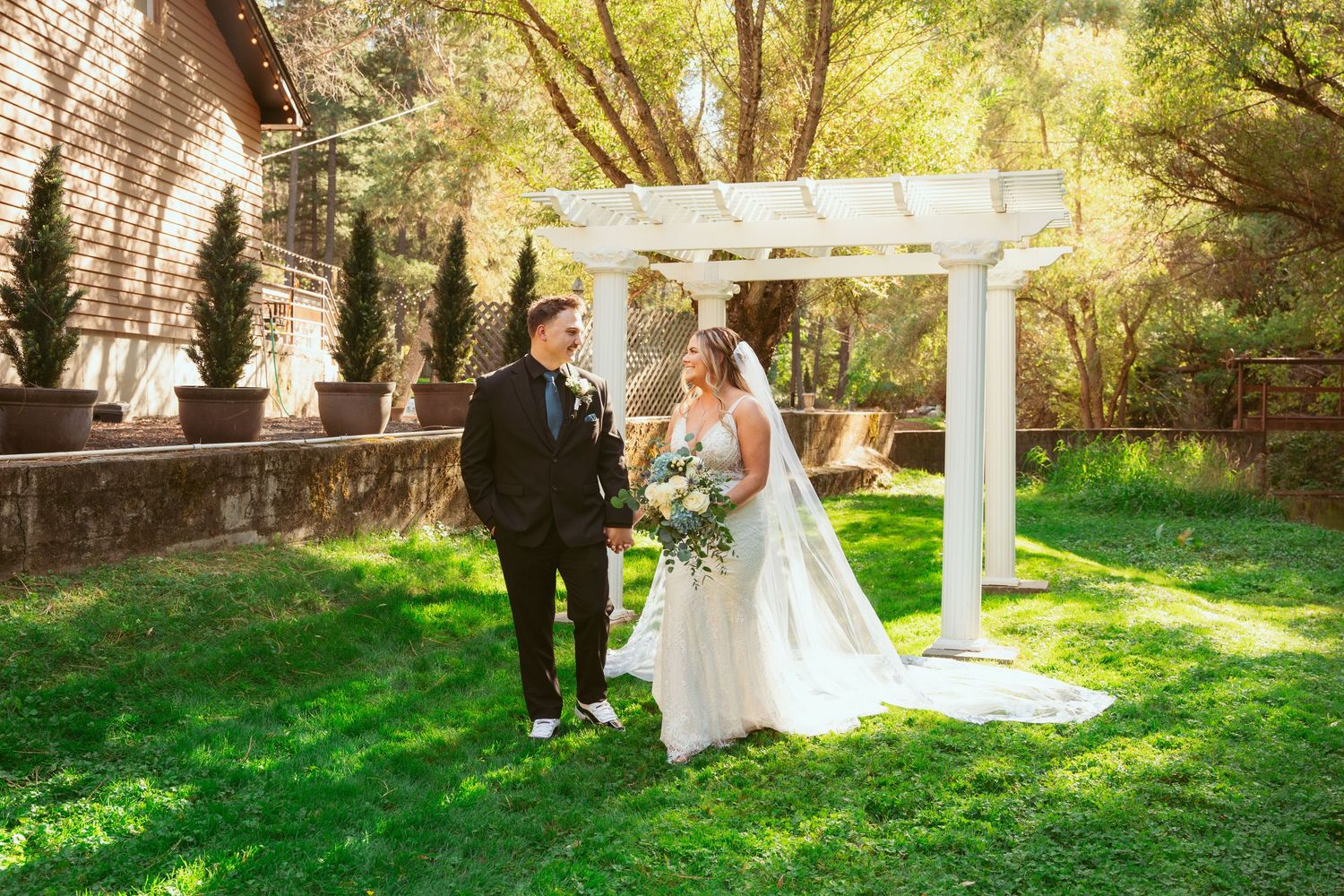 Wedding party poses under a white pergola in a lush garden setting during golden hour.
