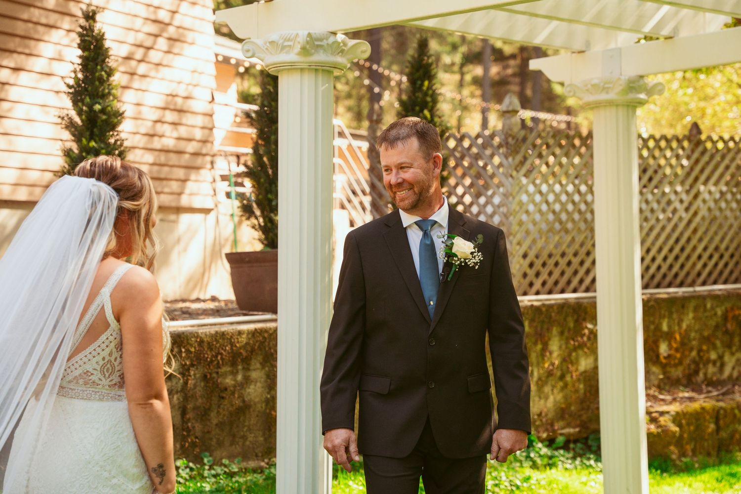 A first look wedding moment captured under a white pergola with bright sunlight filtering through.