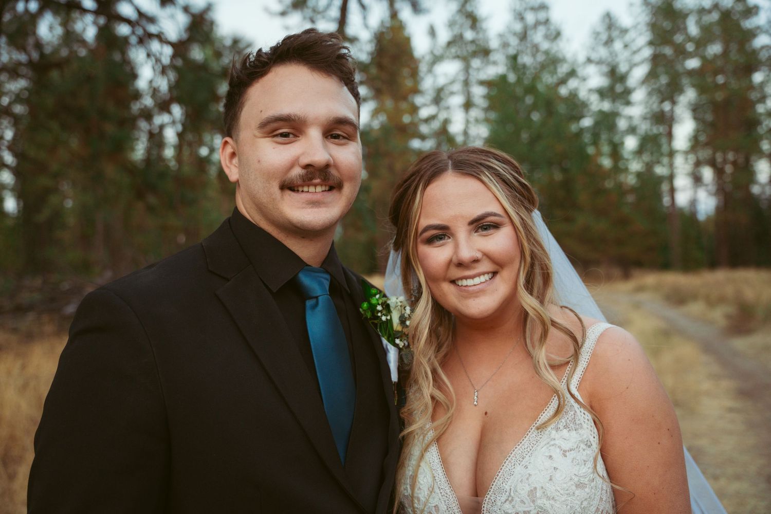 A happy wedding couple poses together outdoors against a backdrop of evergreen trees.