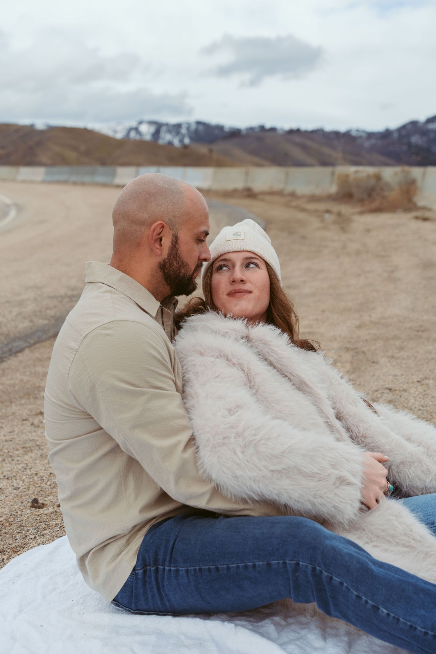 Couple in casual attire cuddling on blanket in desert setting.