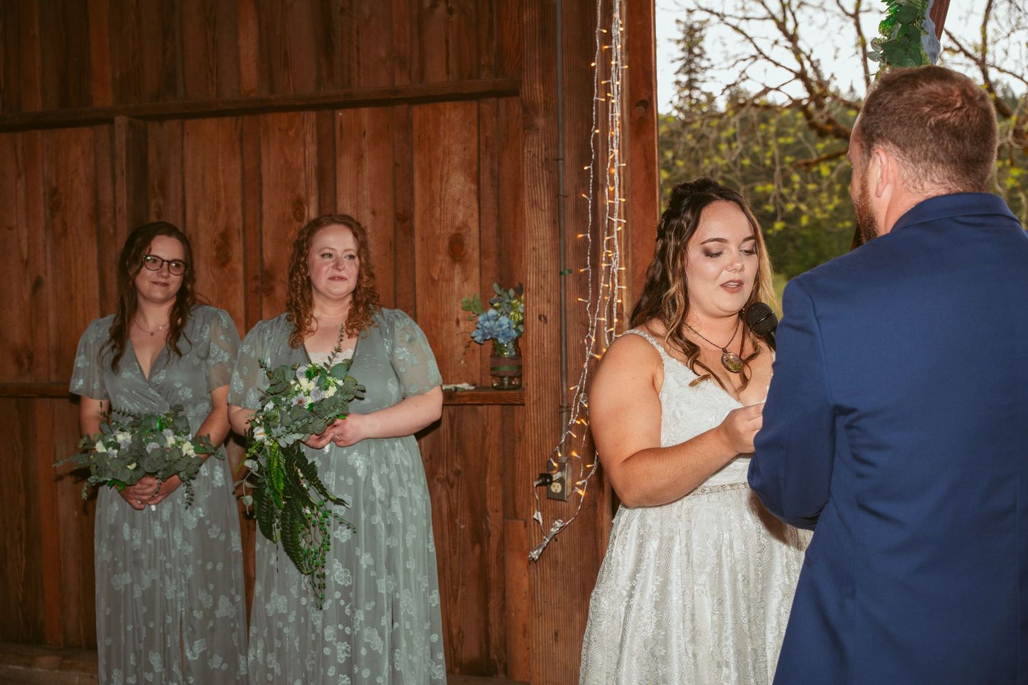 Wedding party in sage green dresses watching ceremony in rustic barn venue.