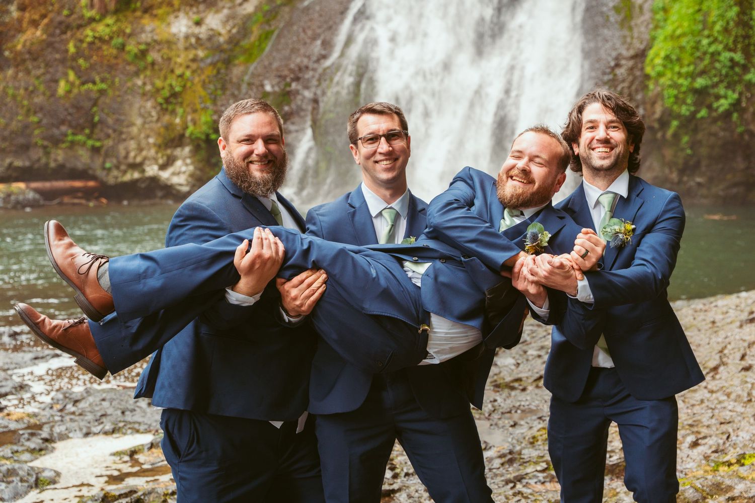 Groomsmen in navy suits pose playfully in front of a scenic waterfall.
