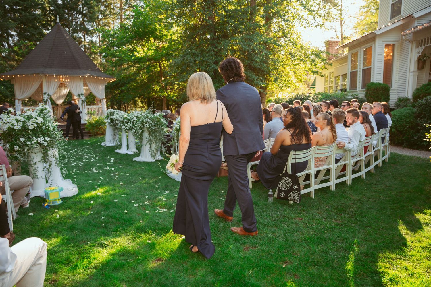Wedding guests gathered on a green lawn during golden hour with white chairs and garden setting.