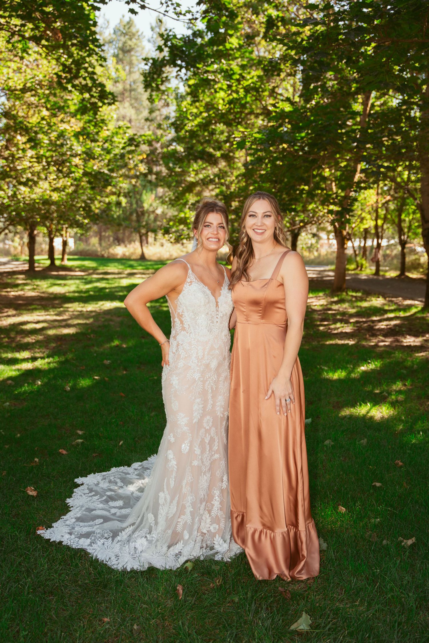 Two women in elegant dresses pose together on a sunny lawn with trees in the background.