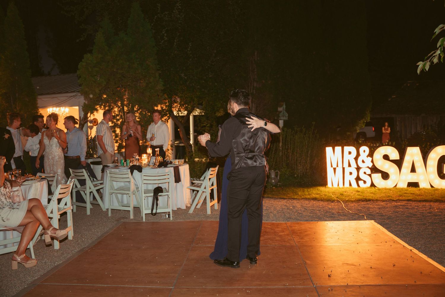Illuminated 'MR & MRS' sign glows at nighttime outdoor wedding reception with dance floor.