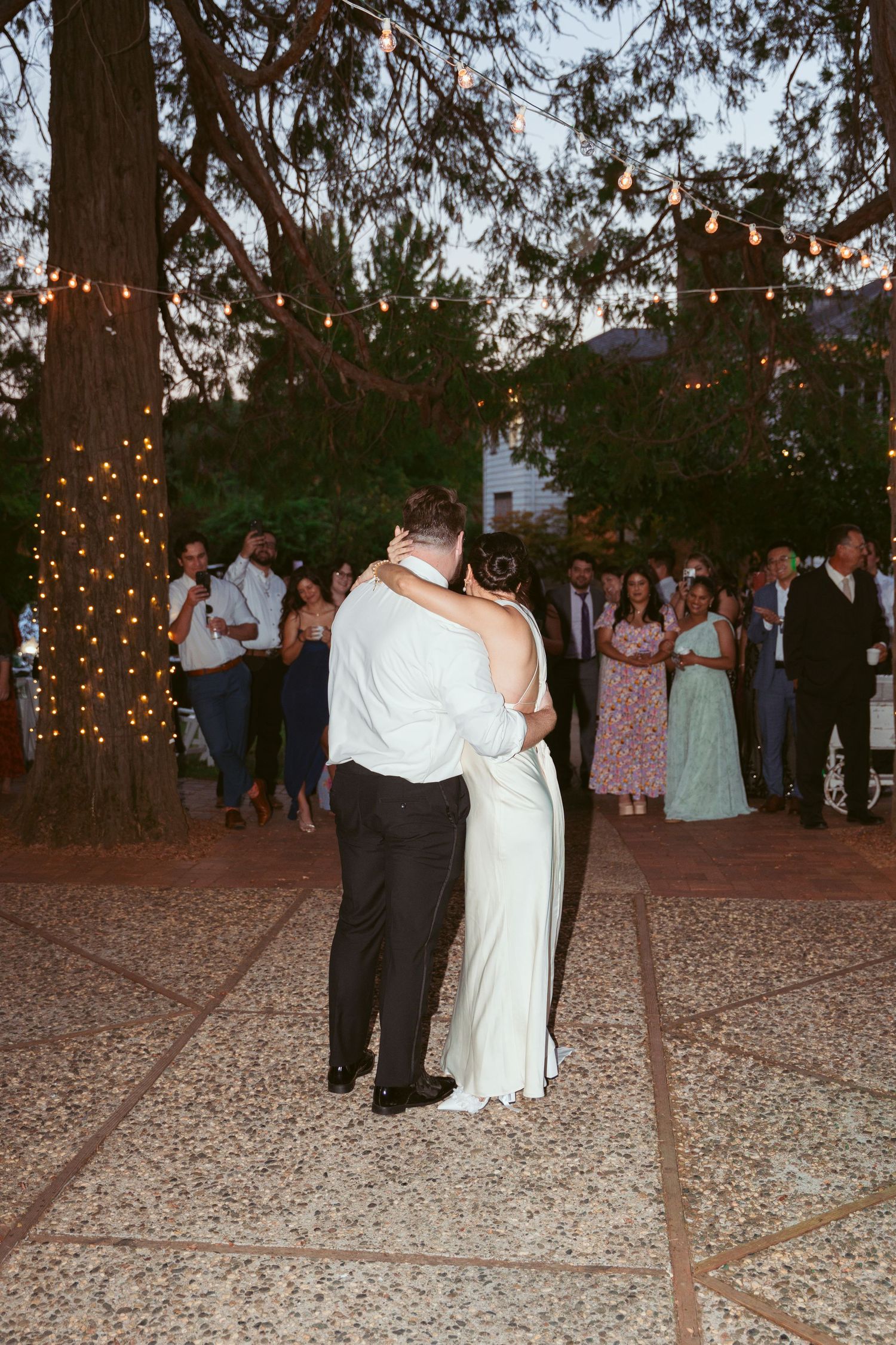 Newlyweds share an intimate dance moment outdoors surrounded by guests and string-lit trees.