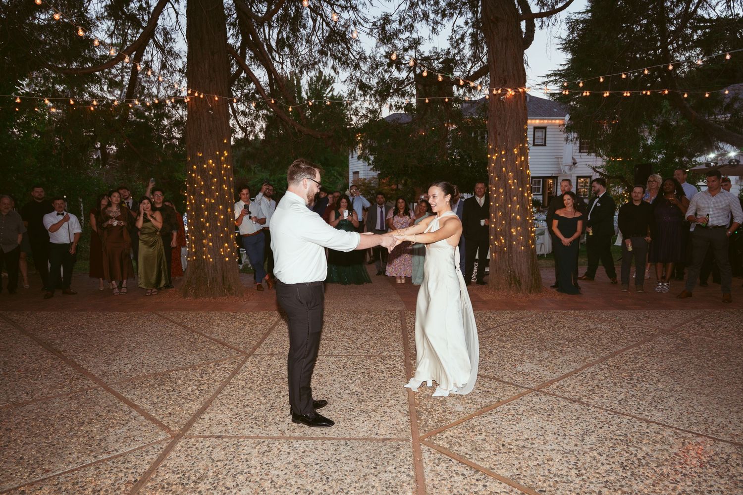 Couples dance together on a rustic outdoor dance floor decorated with string lights at dusk.