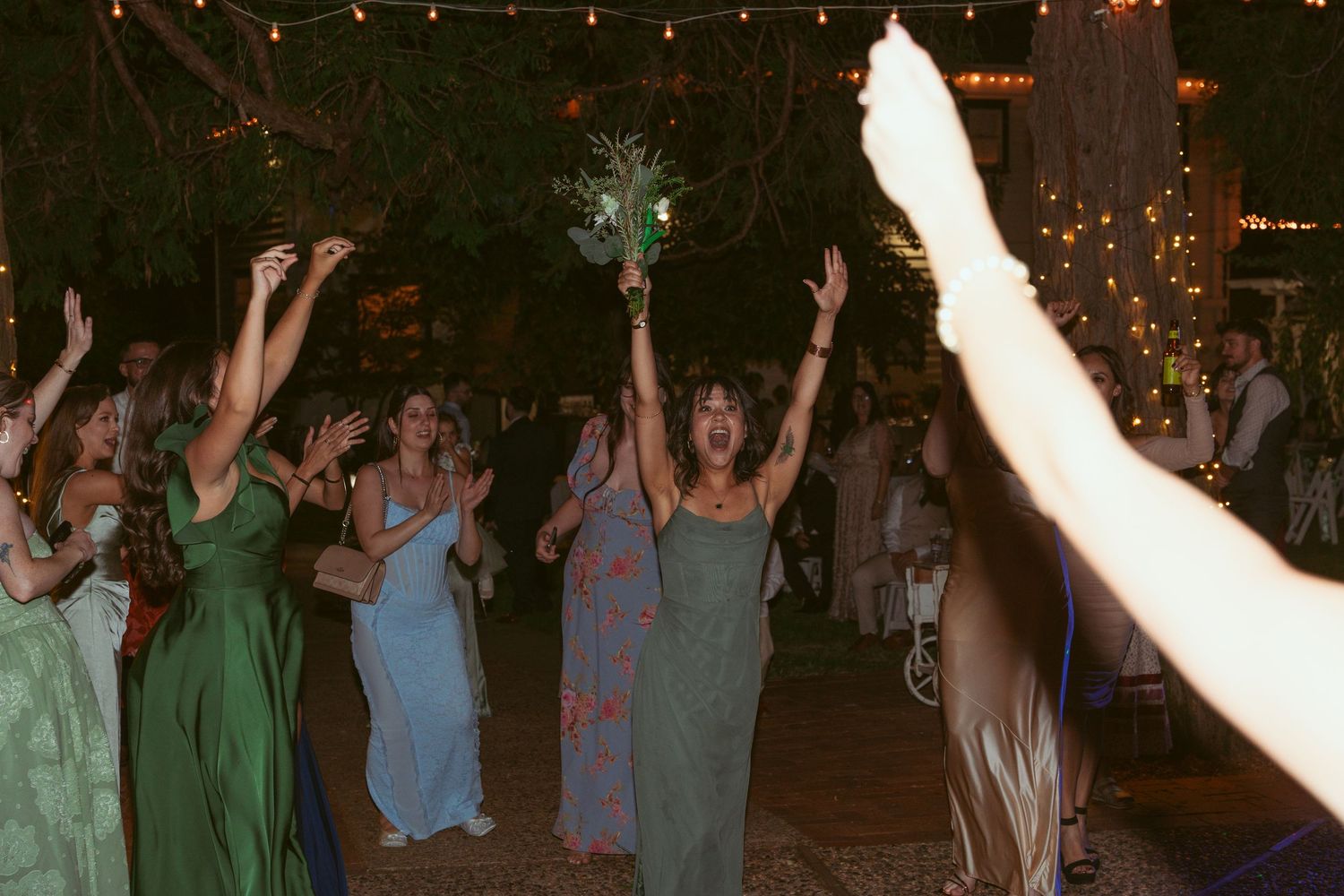 Wedding party and guests dancing with arms raised under twinkling outdoor lights.