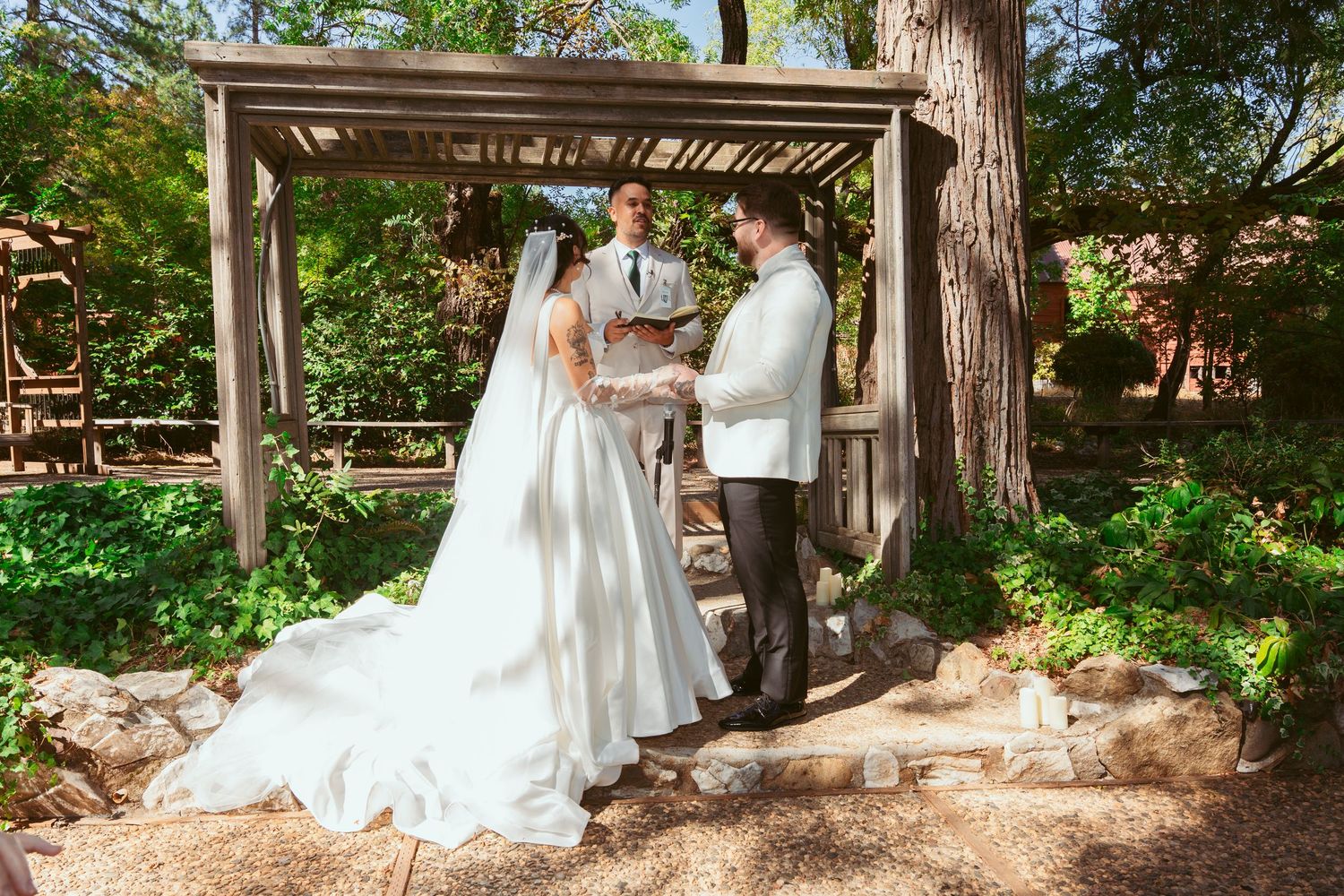 A bride in a white wedding dress poses under a rustic wooden pergola surrounded by lush garden foliage.