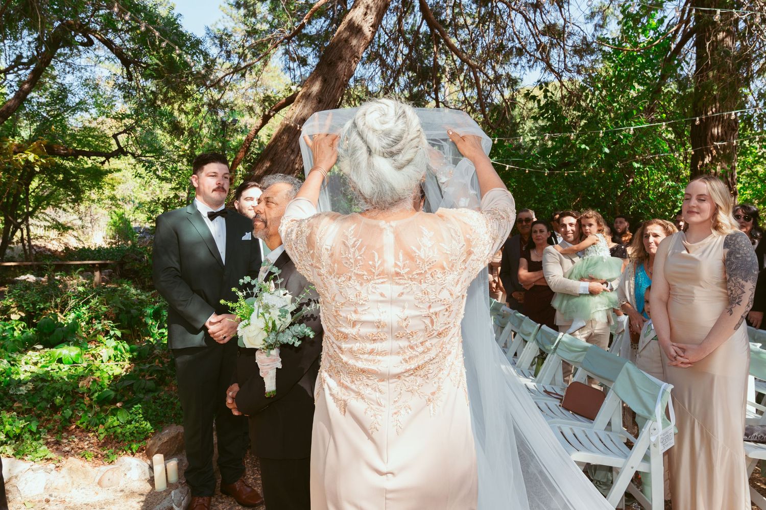 Wedding ceremony guests gather under trees in outdoor garden setting.