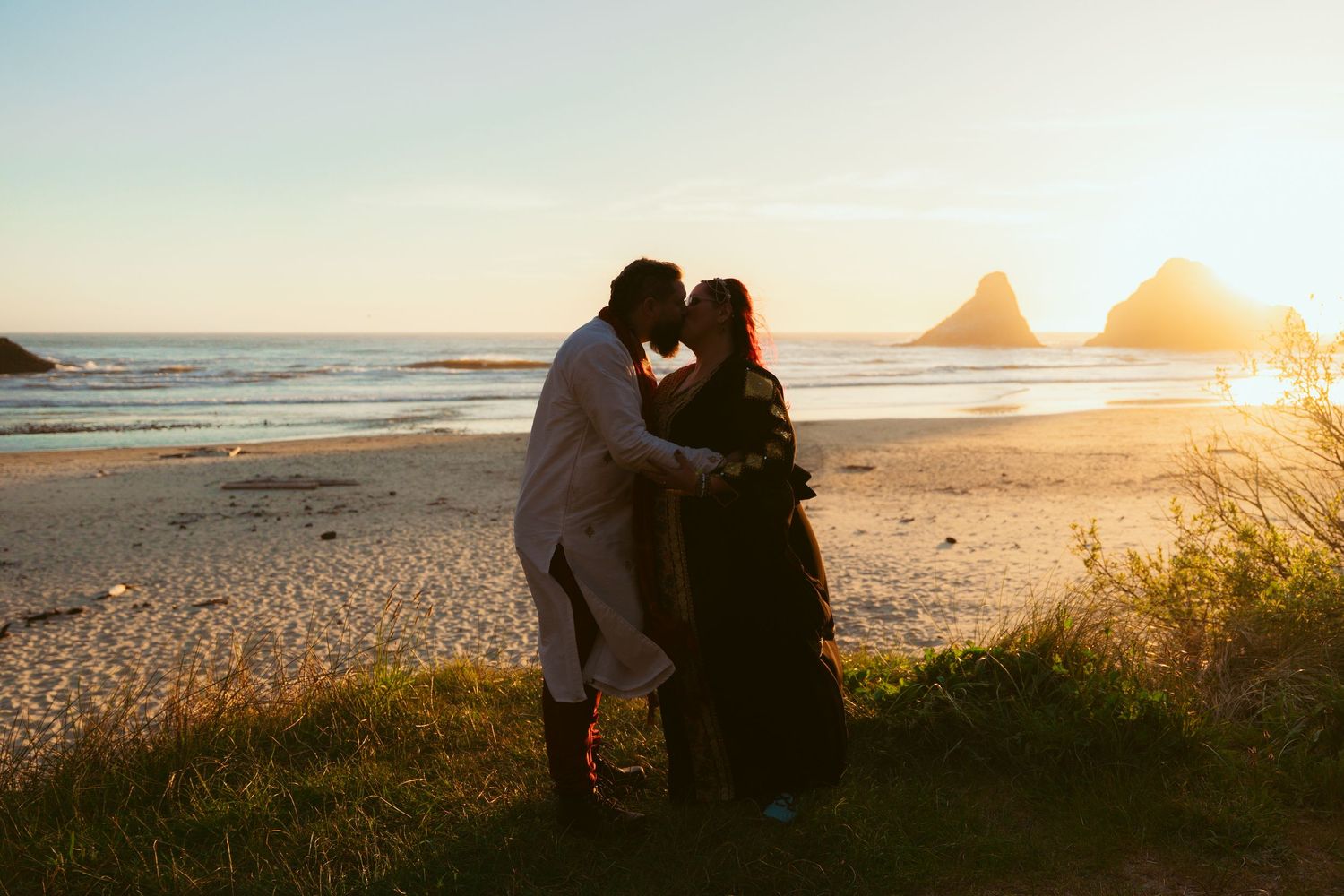 Silhouettes of a couple embracing against a golden sunset at the beach with sea stacks visible.