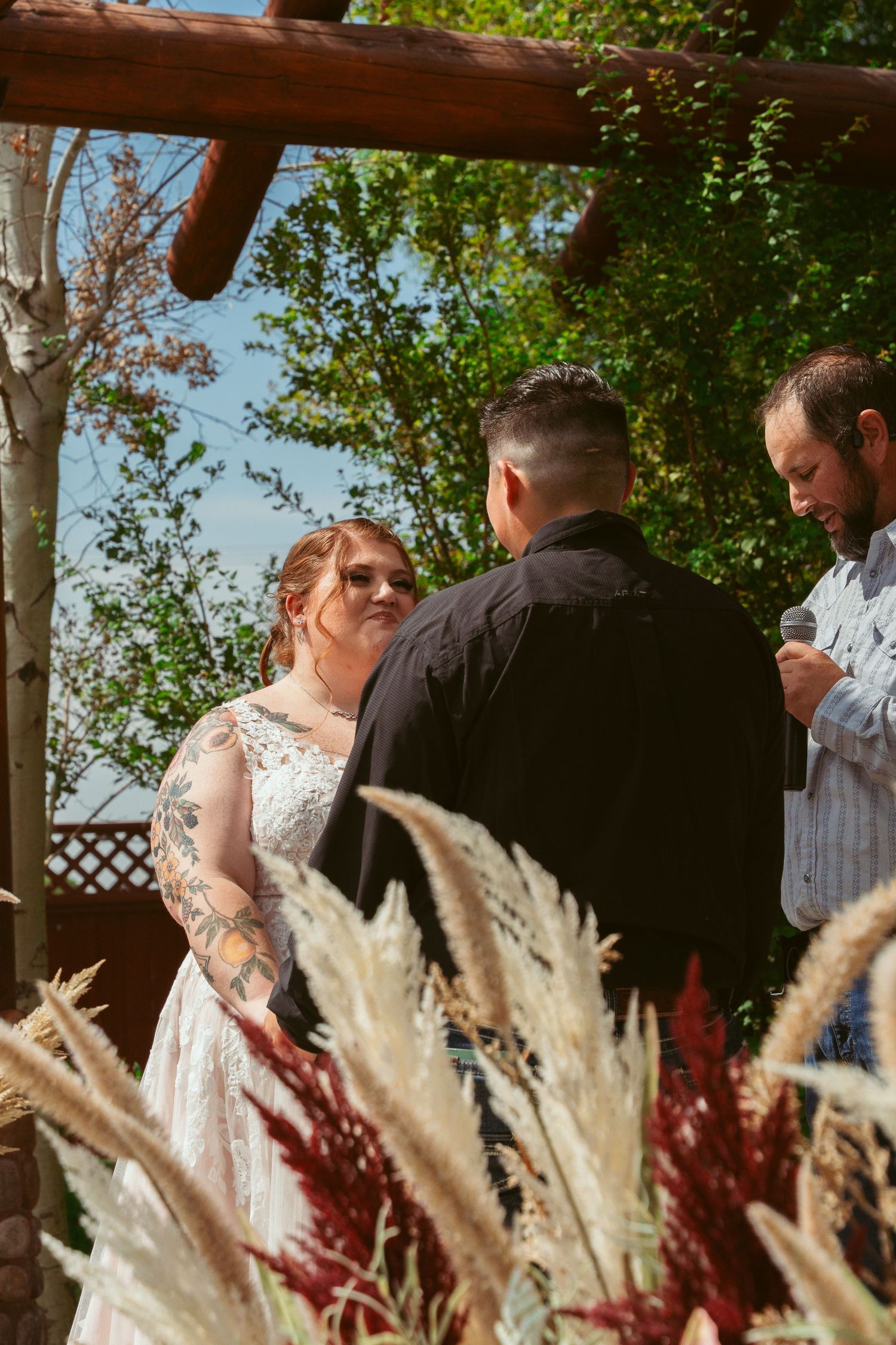 Wedding ceremony under a rustic wooden pergola with decorative pampas grass in the foreground.
