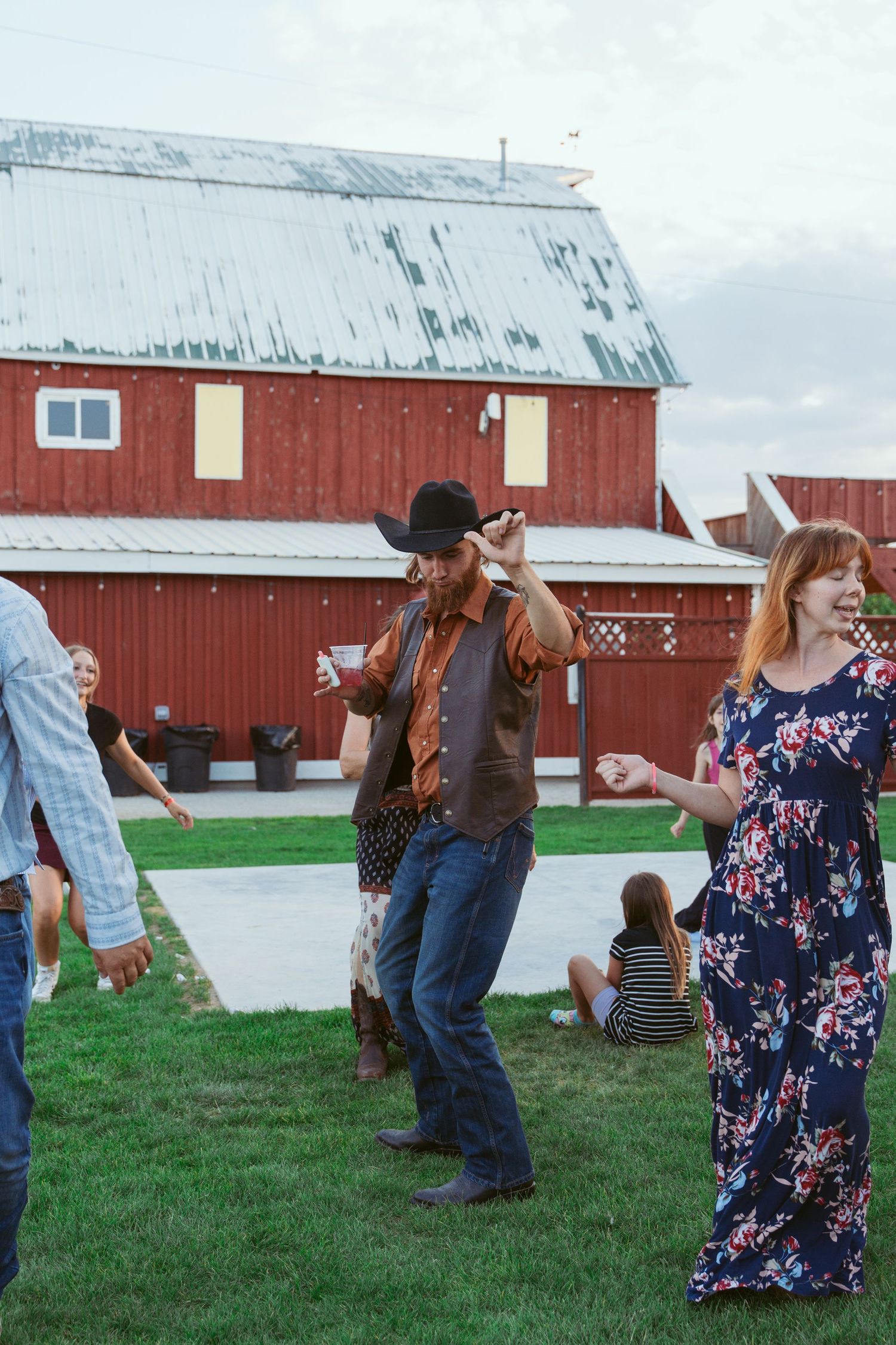 People dancing and celebrating at an outdoor party near a red barn on a grassy lawn.
