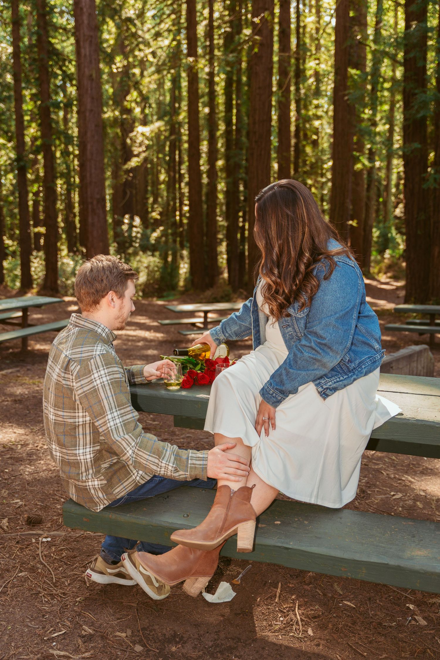 Two people share a playful moment on a wooden bench in a redwood forest setting.