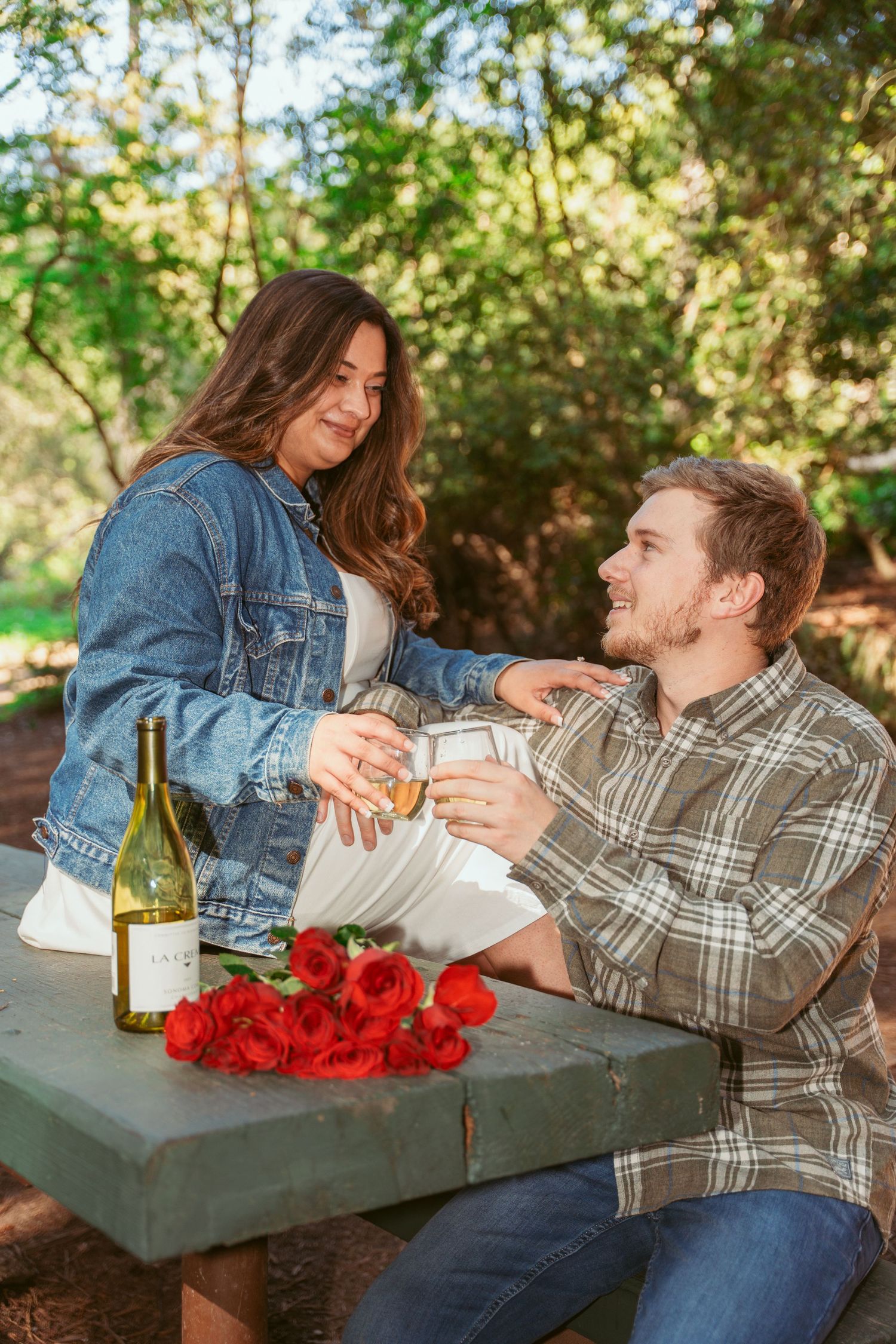 Couple sharing wine and roses at outdoor picnic table in sunlit forest.