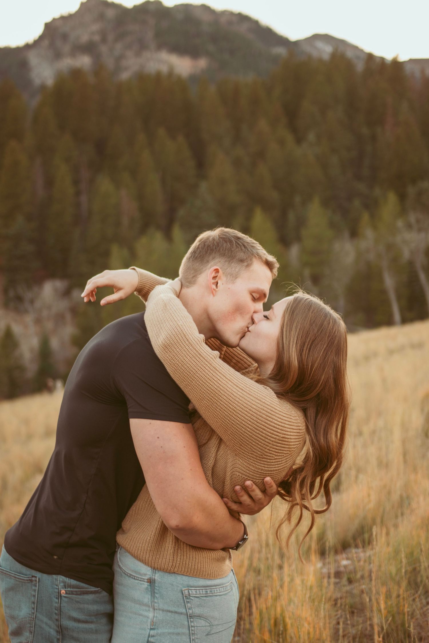 Romantic embrace in golden mountain meadow with evergreen forest backdrop.