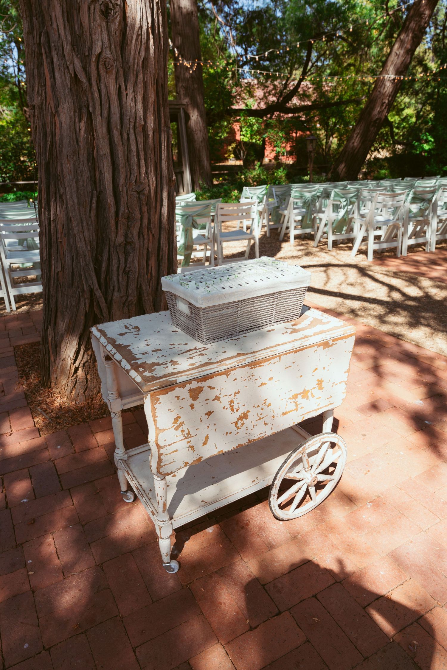 Vintage white wooden cart with wheels displayed in a garden setting with large trees and dappled sunlight.