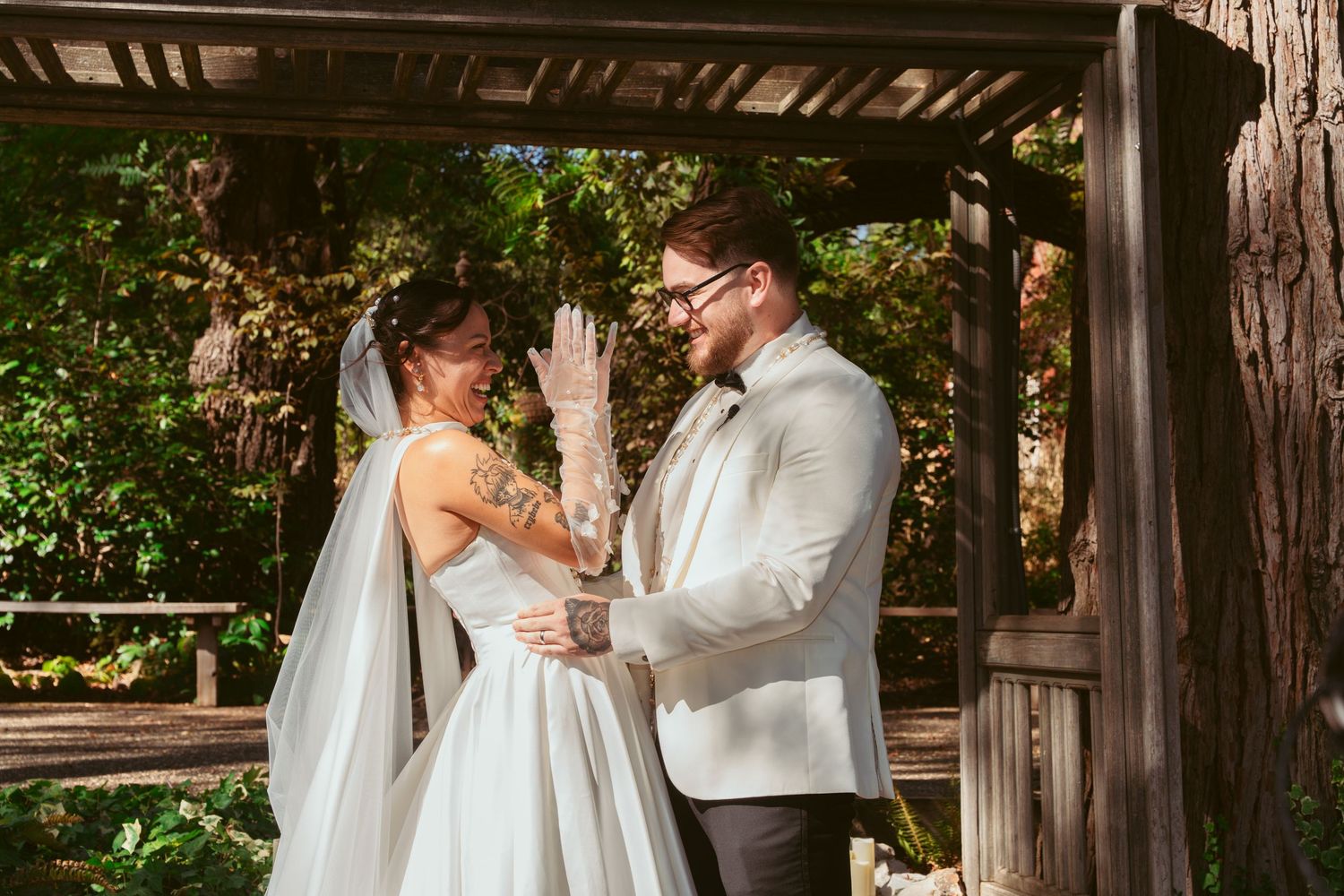 Wedding couple exchanges vows under wooden beams with sunlight filtering through trees.