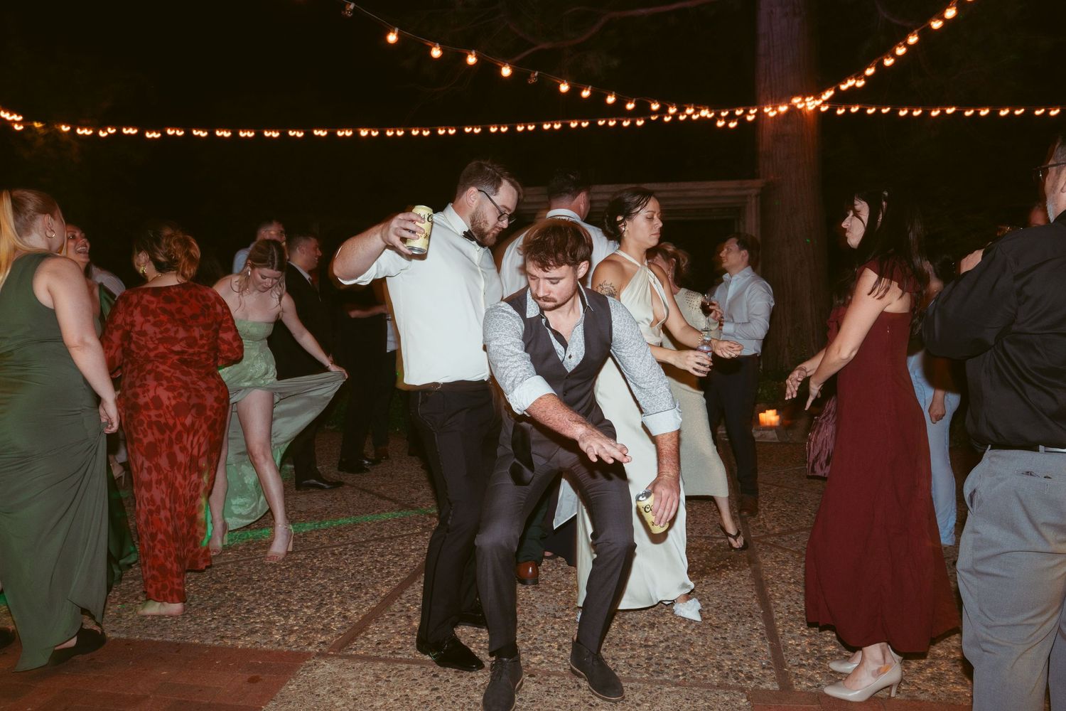 Wedding guests dance enthusiastically under string lights at an outdoor evening reception.