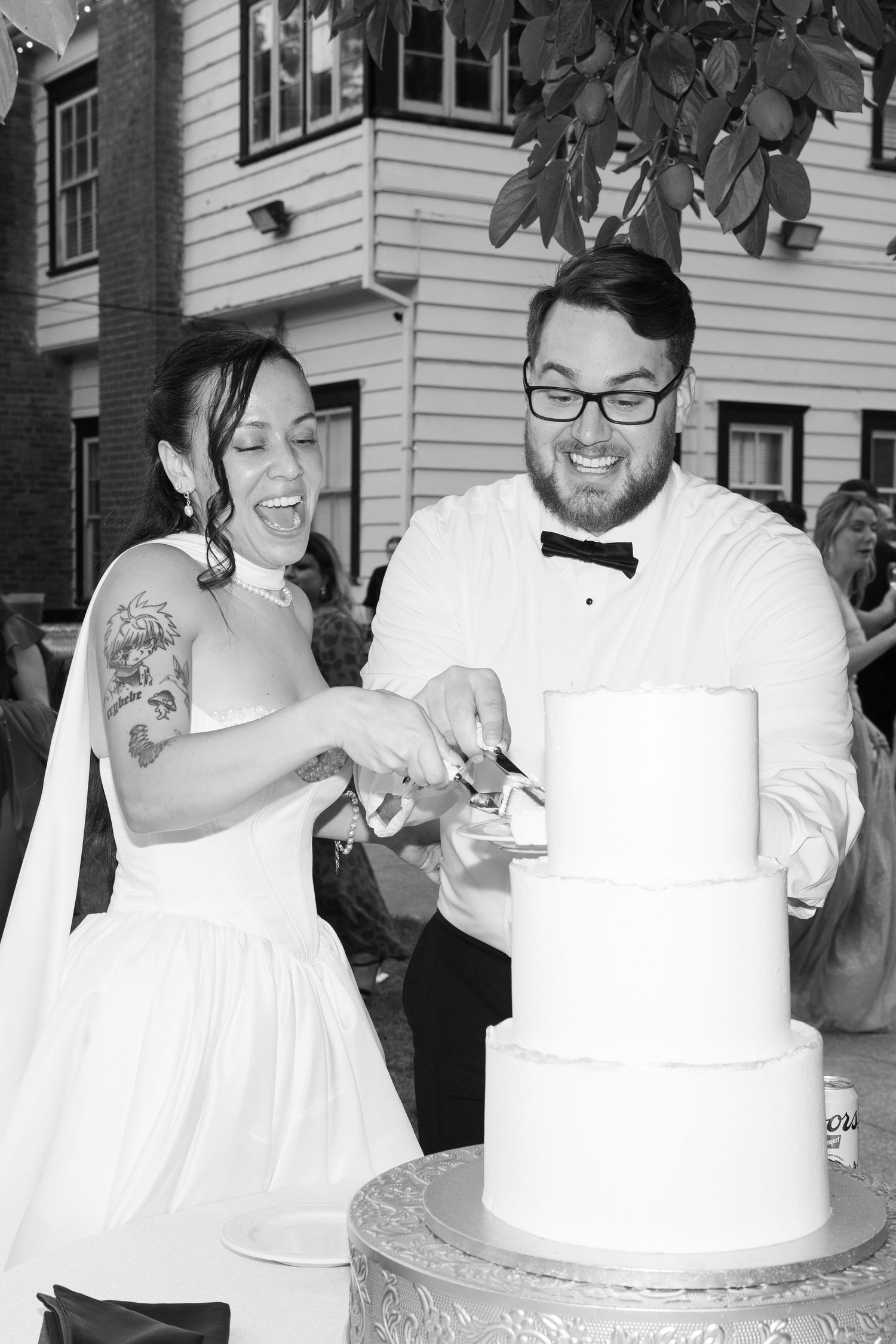 A couple shares a playful moment while cutting their white wedding cake at an outdoor reception.
